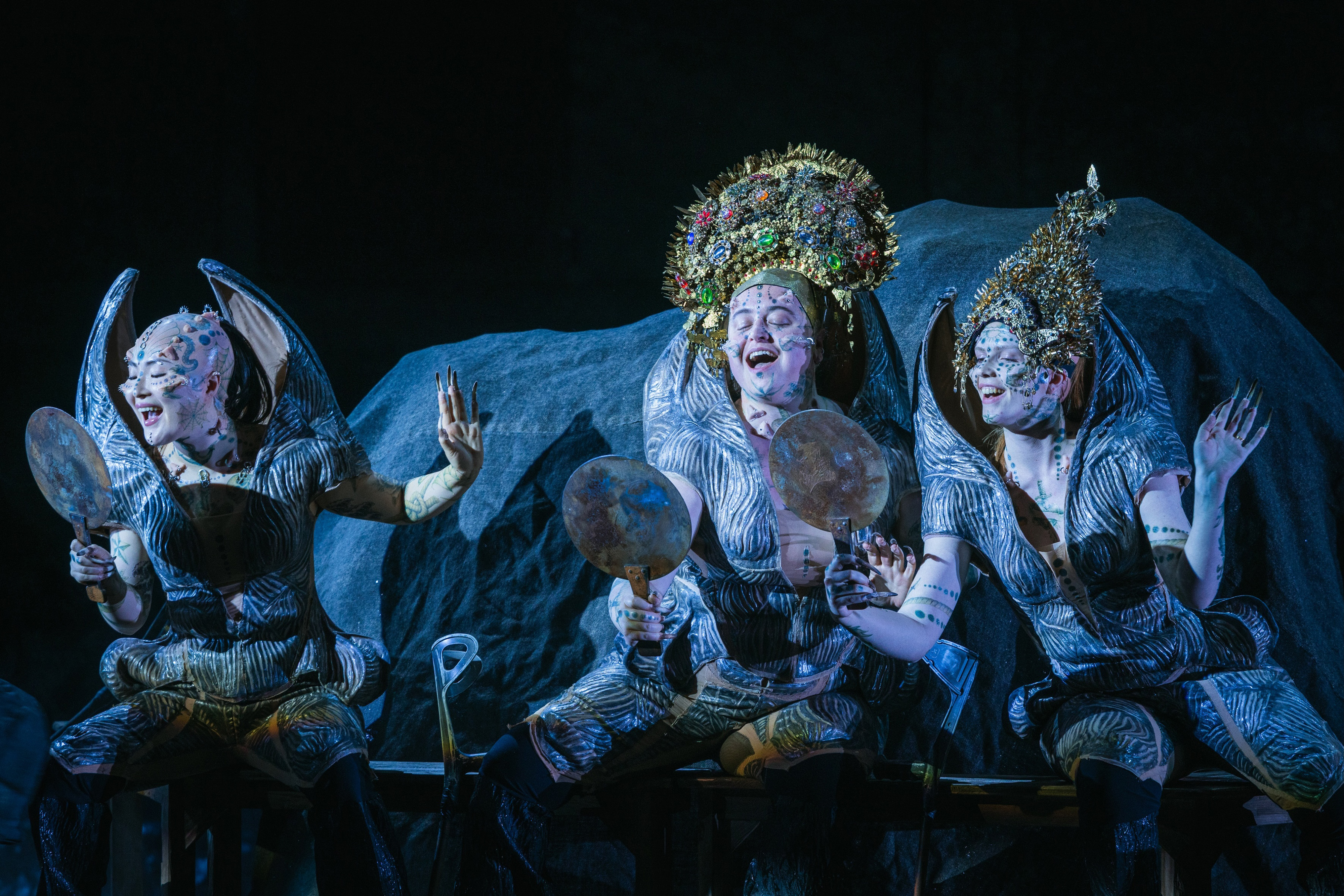 Three performers in elaborate costumes and ornate headdresses sit onstage, holding round mirrors and gesturing expressively. The background features a large rock-like structure.