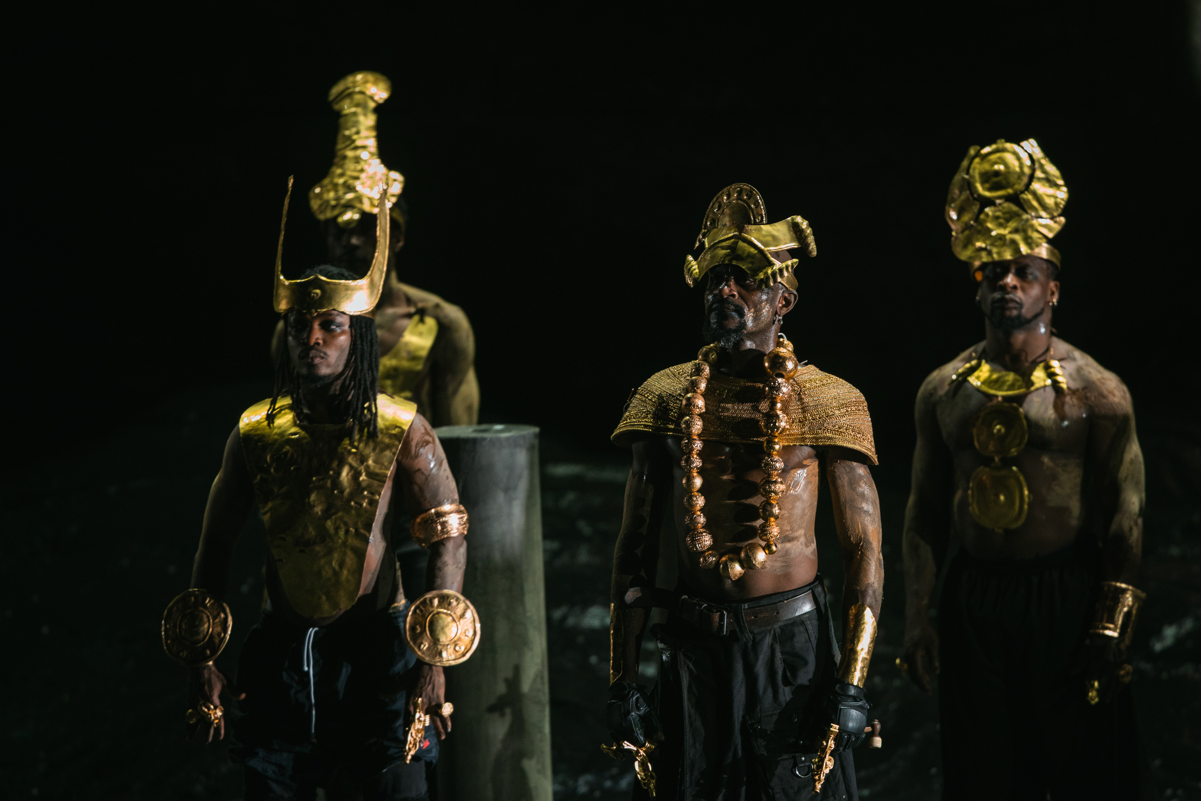 Vier Männer stehen in schummrigem Licht, hemdsärmelig, mit aufwendigen goldenen Kopfbedeckungen, Schmuck und Brustpanzern, und blicken mit ernster Miene nach vorne. Der Hintergrund ist dunkel und schattig.