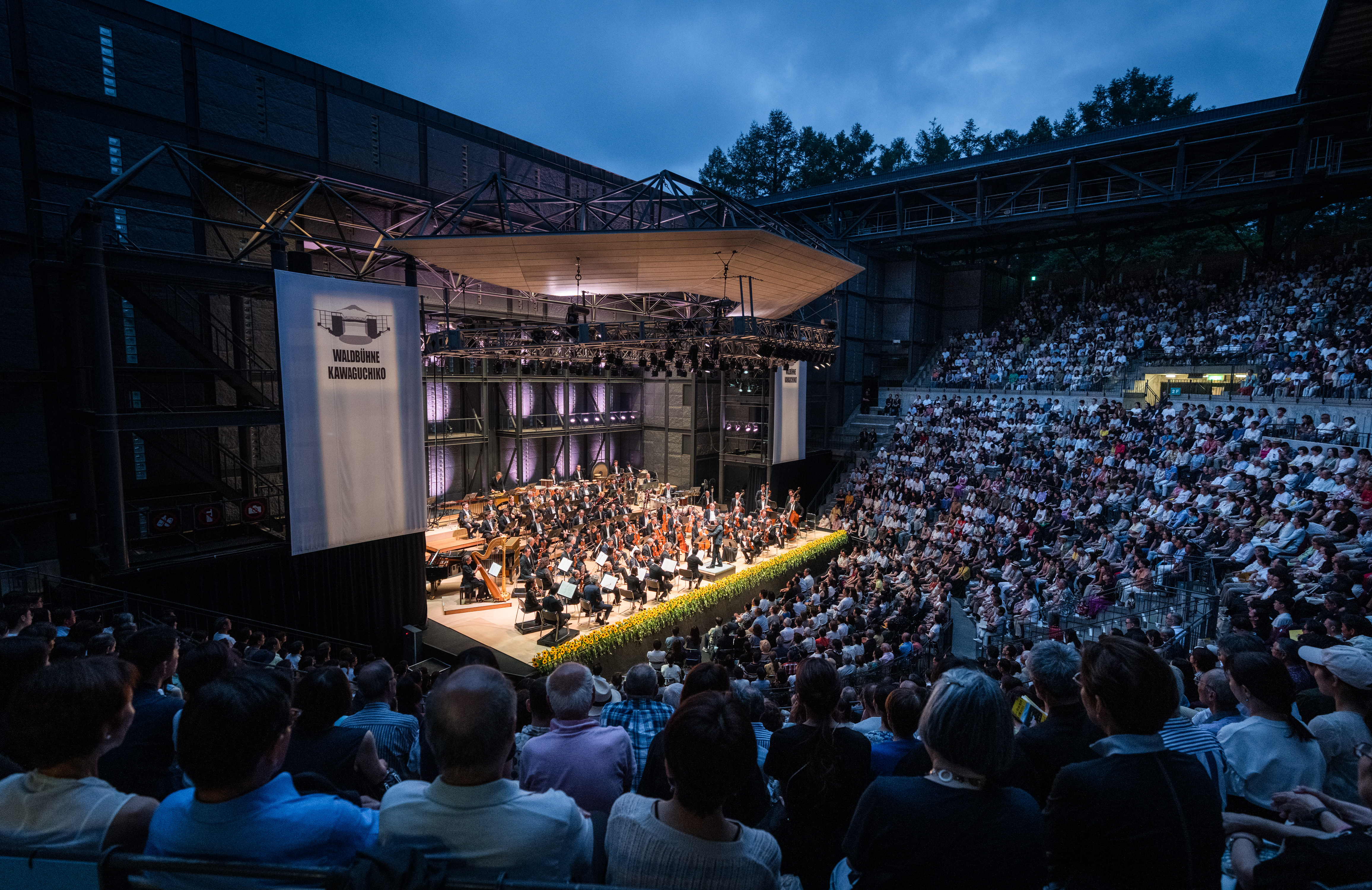 Das Amphitheater in der japanischen Stadt Kawaguchiko unter freiem Himmel, das mit Menschen gefüllt ist, die ein Orchester auf einer großen, gut beleuchteten Bühne unter einem Vordach spielen sehen, wobei die Bäume in der Abenddämmerung im Hintergrund zu sehen sind.