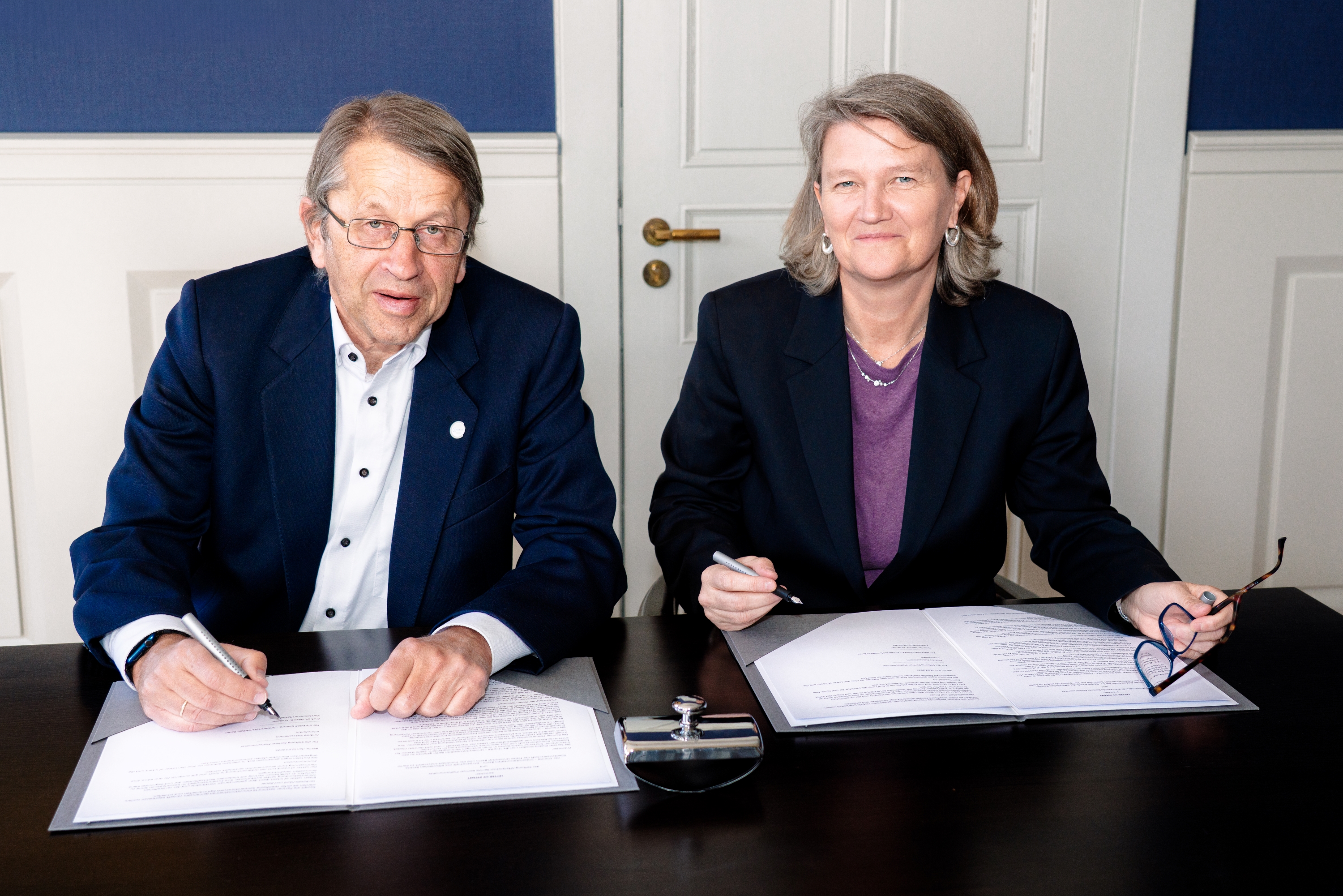 Prof. Heyo K. Kroemer und Andrea Zietzschmann in Geschäftskleidung sitzen an einem Tisch und unterschreiben jeweils ein Dokument. Beide lächeln und schauen in die Kamera. Auf dem Tisch vor ihnen liegen Papiere, ein Stiftständer und eine Brille.