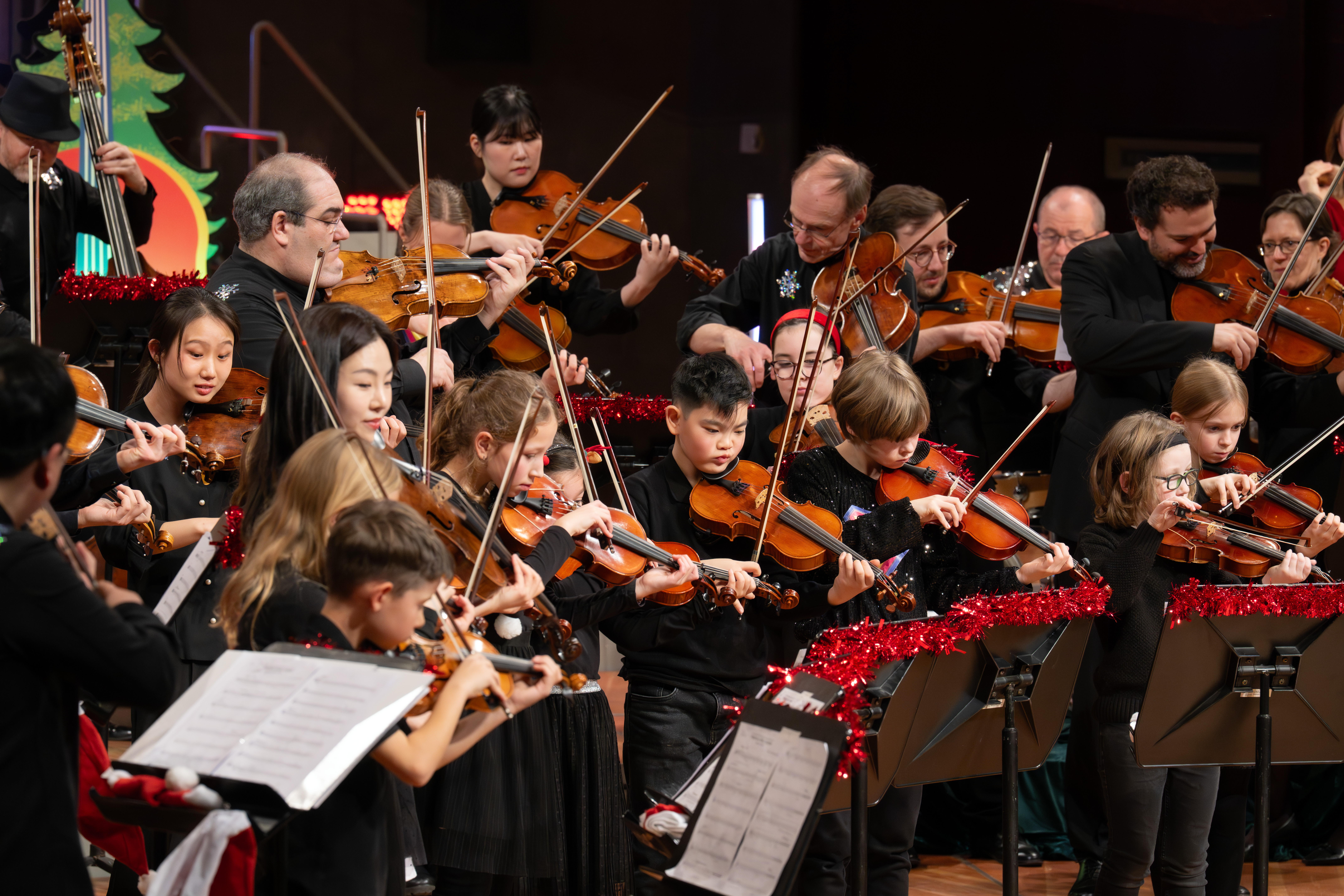 Schwarz gekleidete Kinder und Erwachsene spielen gemeinsam in einem Orchester Geige. Rotes Lametta schmückt die Notenständer, und im Hintergrund ist ein Weihnachtsbaum zu sehen, der auf eine festliche Aufführung hindeutet.