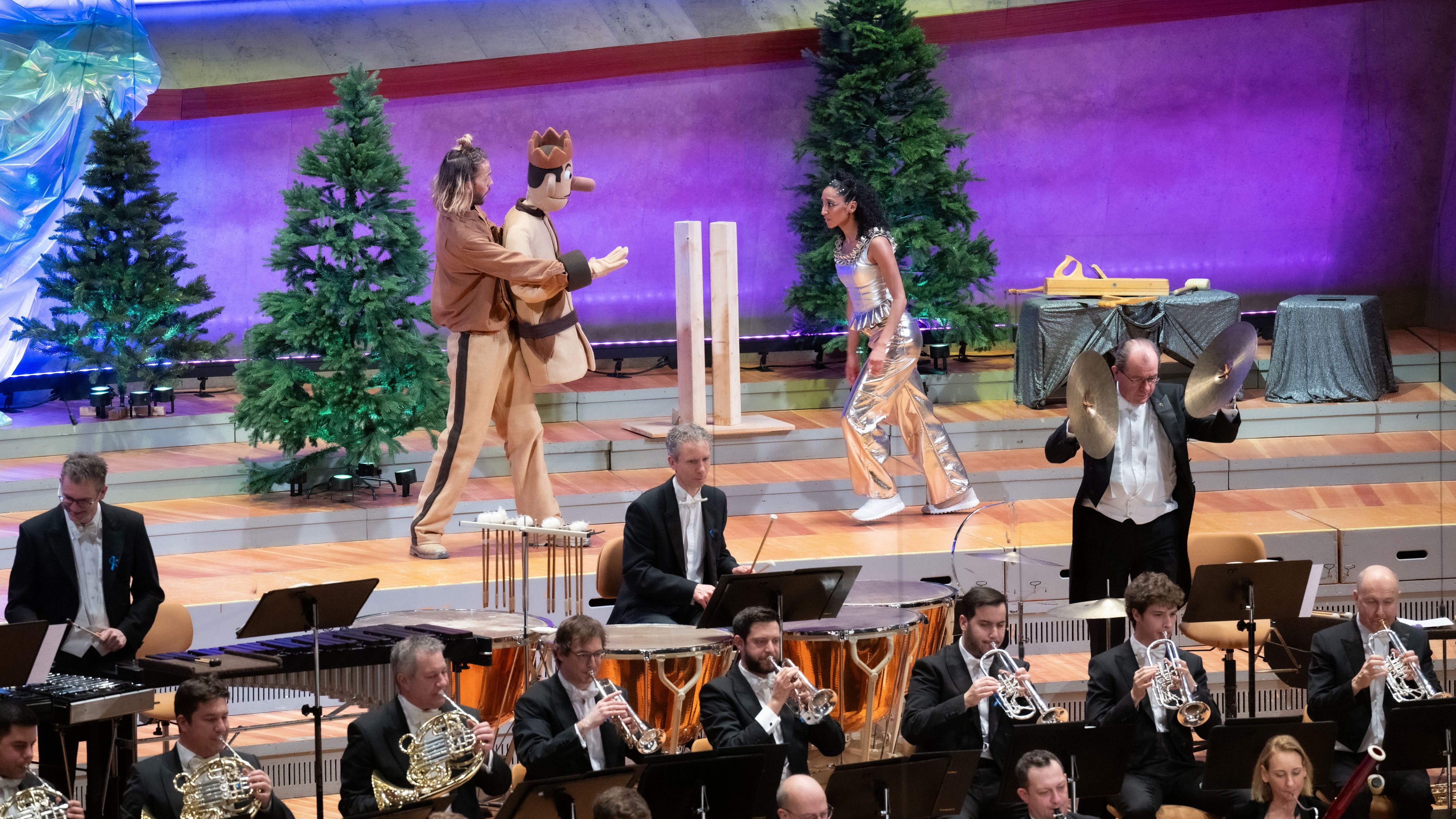 At the front, part of the orchestra can be seen on stage, at the back a man with a large prince puppet walks towards a dressed up woman