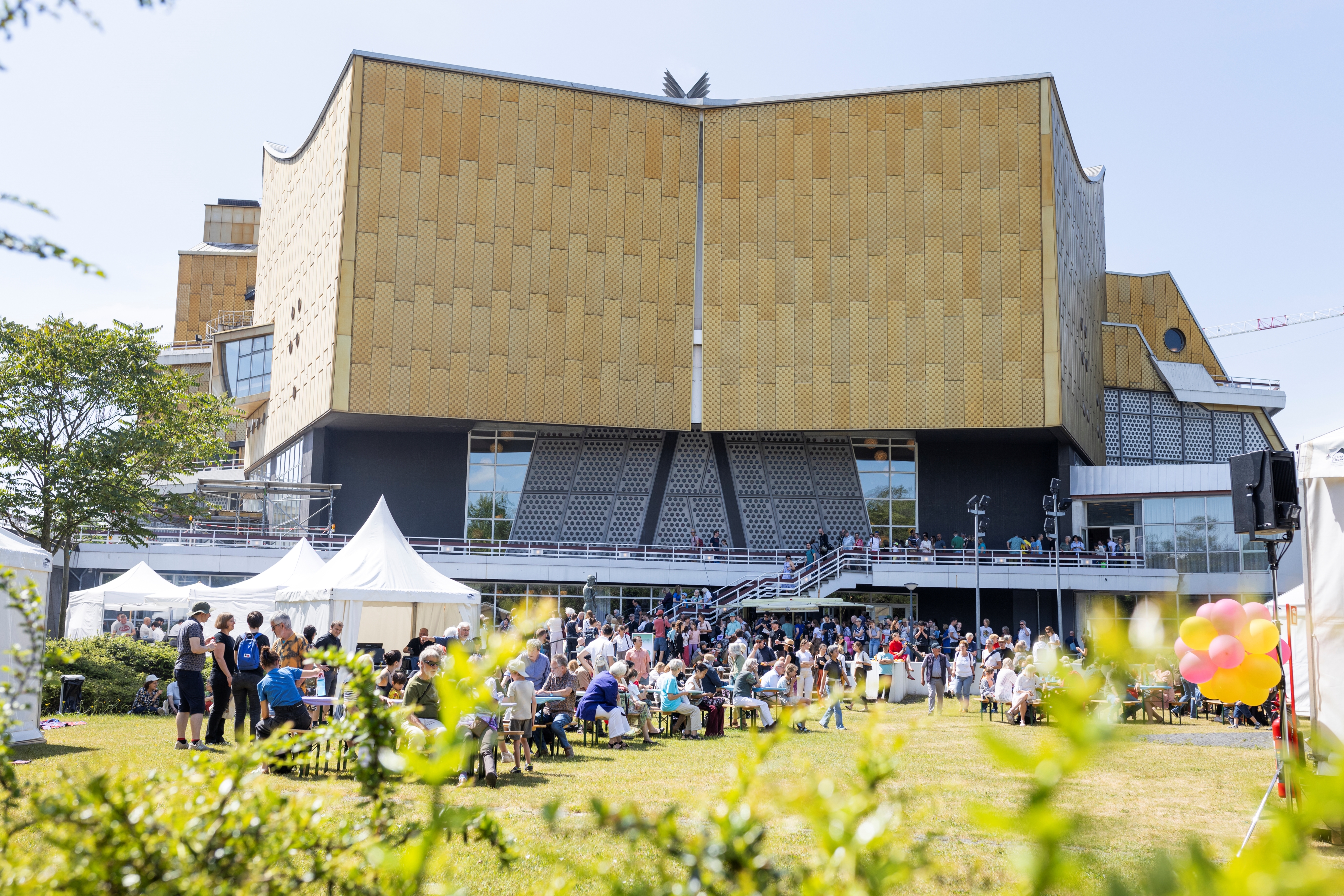 Eine große Menschenmenge versammelt sich vor einem modernen, goldfarbenen Gebäude mit einzigartigem architektonischen Design. An diesem sonnigen Tag sitzen und stehen die Menschen, Zelte und bunte Luftballons tragen zur festlichen Atmosphäre bei.
