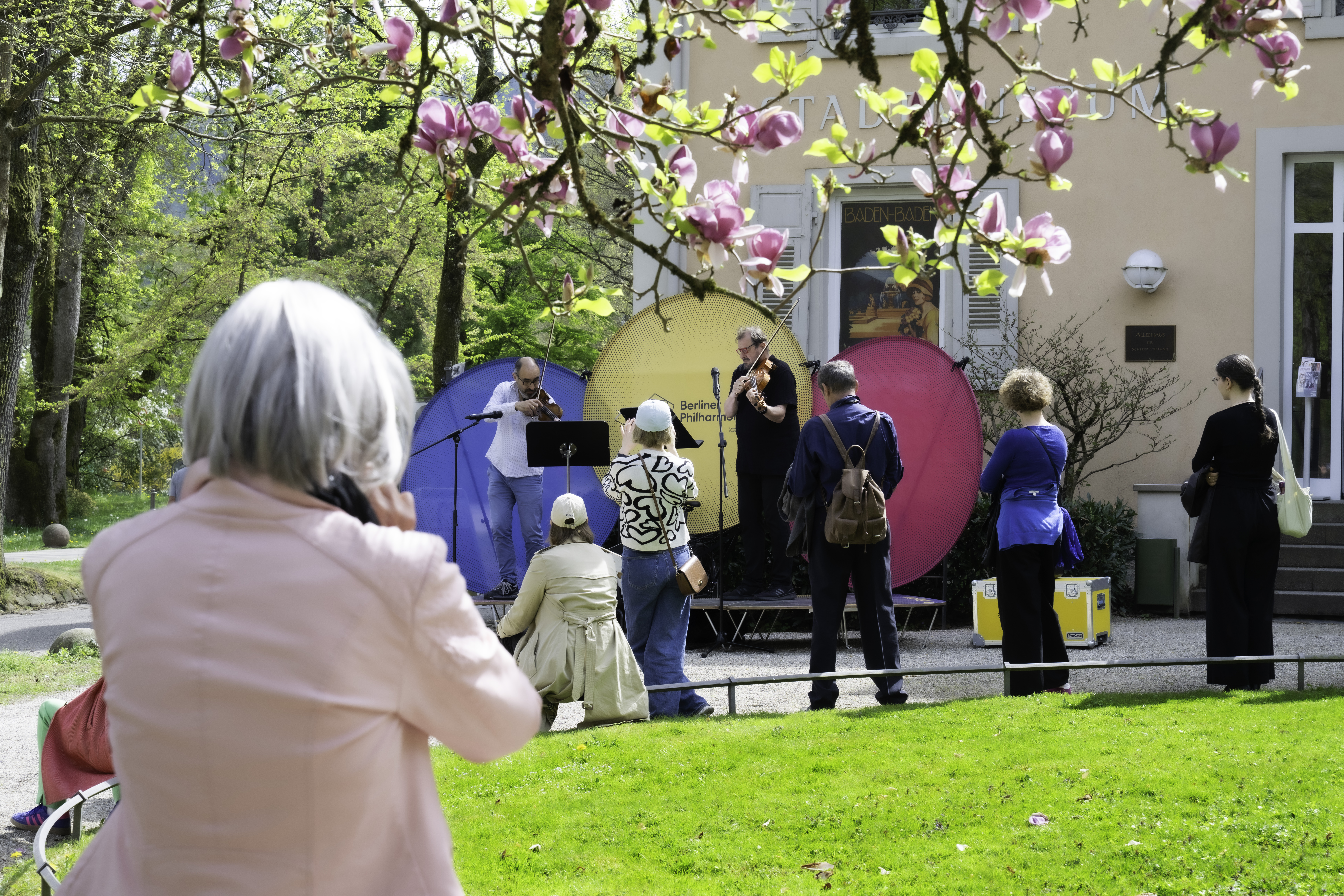 Eine Gruppe von Menschen sieht sich eine kleine Musikaufführung im Freien unter blühenden rosa Magnolienbäumen an; eine Frau im Vordergrund telefoniert, andere stehen oder sitzen im Gras und schauen den Musikern zu.