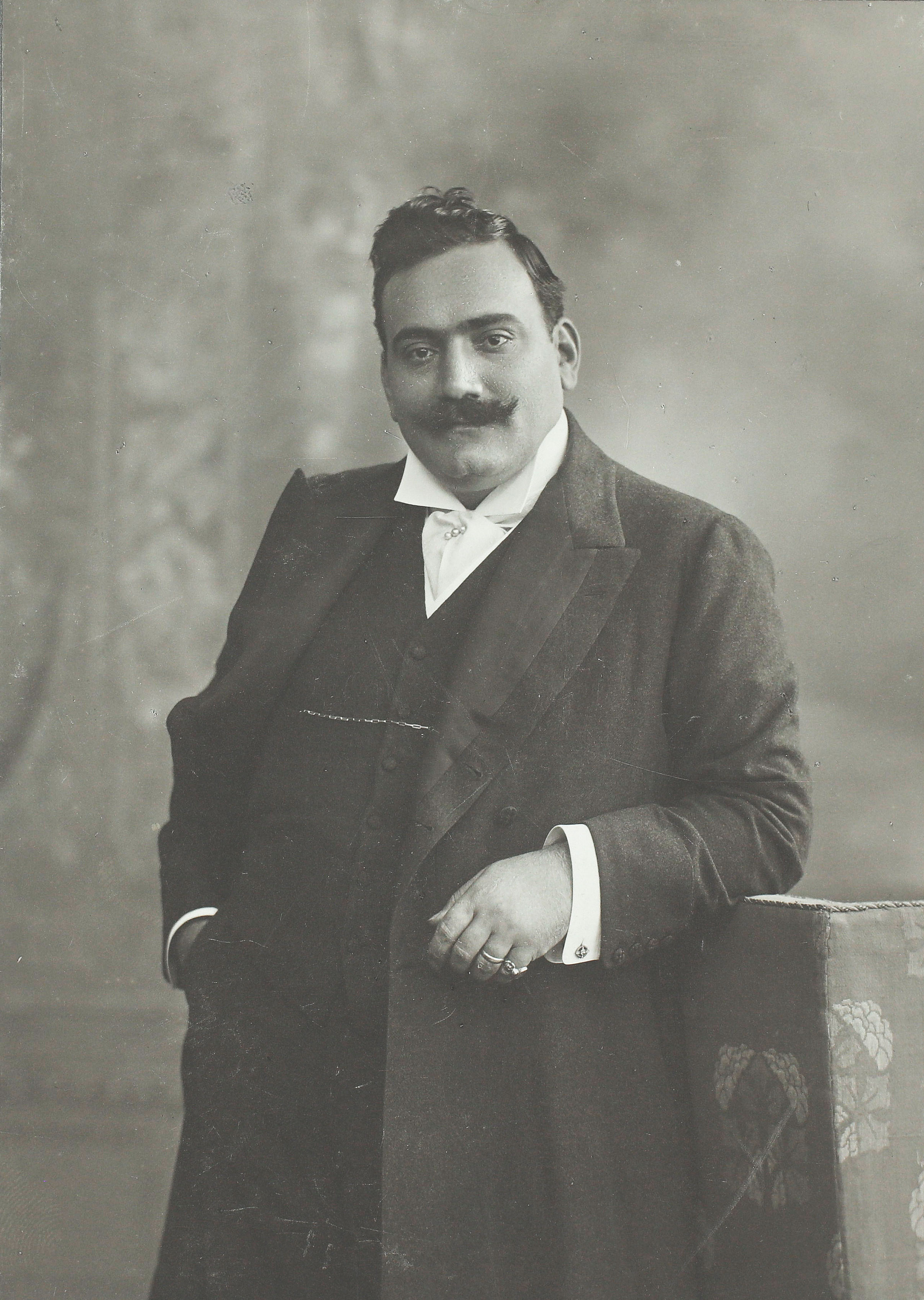 Ein Mann mit Schnurrbart in einem formellen Anzug posiert für ein klassisches Schwarz-Weiß-Porträt, wobei er eine Hand in der Tasche hat und die andere auf einer gemusterten Oberfläche ruht.