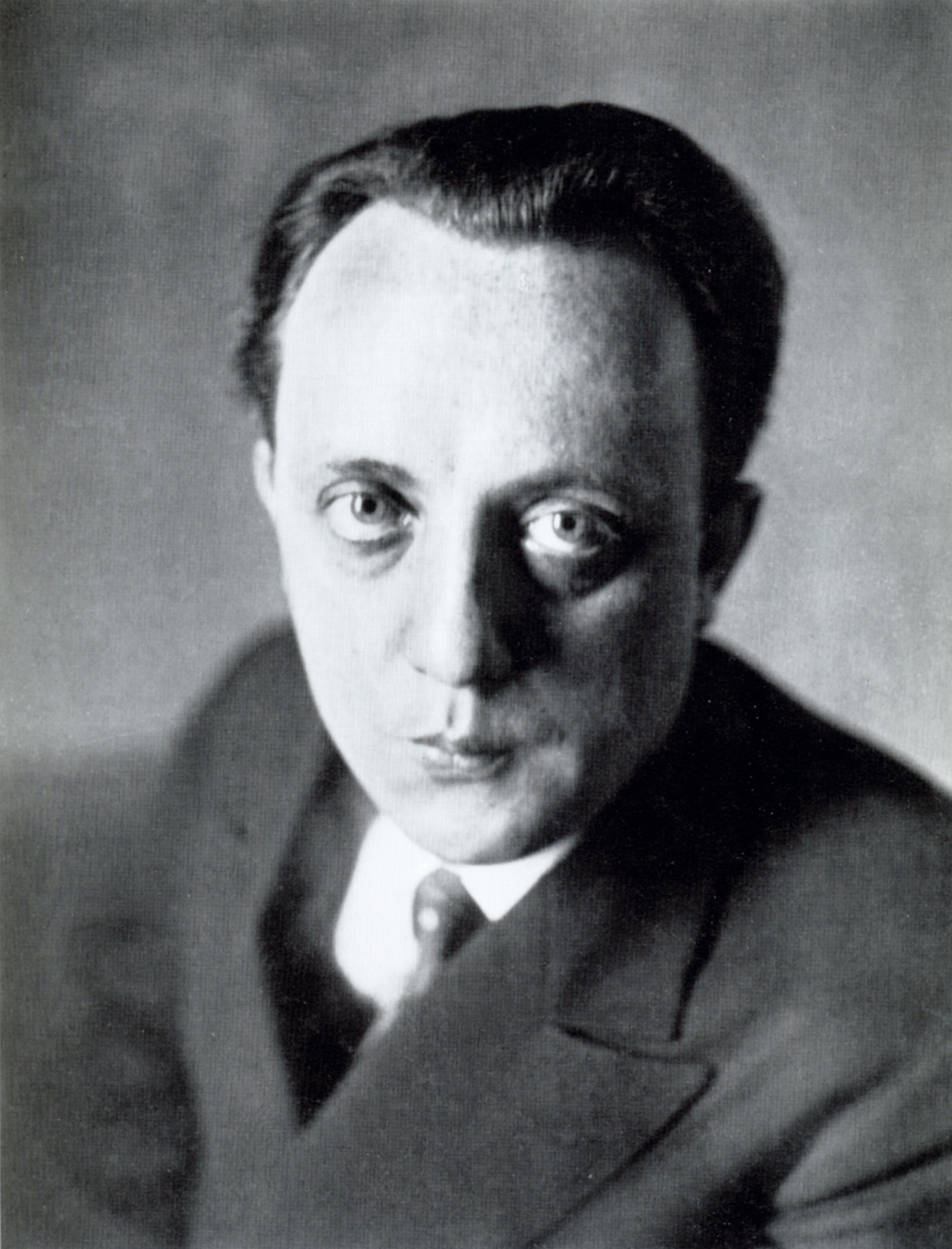 Schwarz-Weiß-Fotographie eines Mannes, der ein dunkles Sakko trägt mit weißem Hemd und Krawatte. Sein Kopf ist frontal nach vorne gerichtet, während sein Oberkörper mit einer Schulter leicht nach hinten gedreht ist. Seine großen Augen blicken direkt in die Kamera, sein Mund ist geschlossen. Er hat dunkle Haare und eine Kurzhaarfrisur.