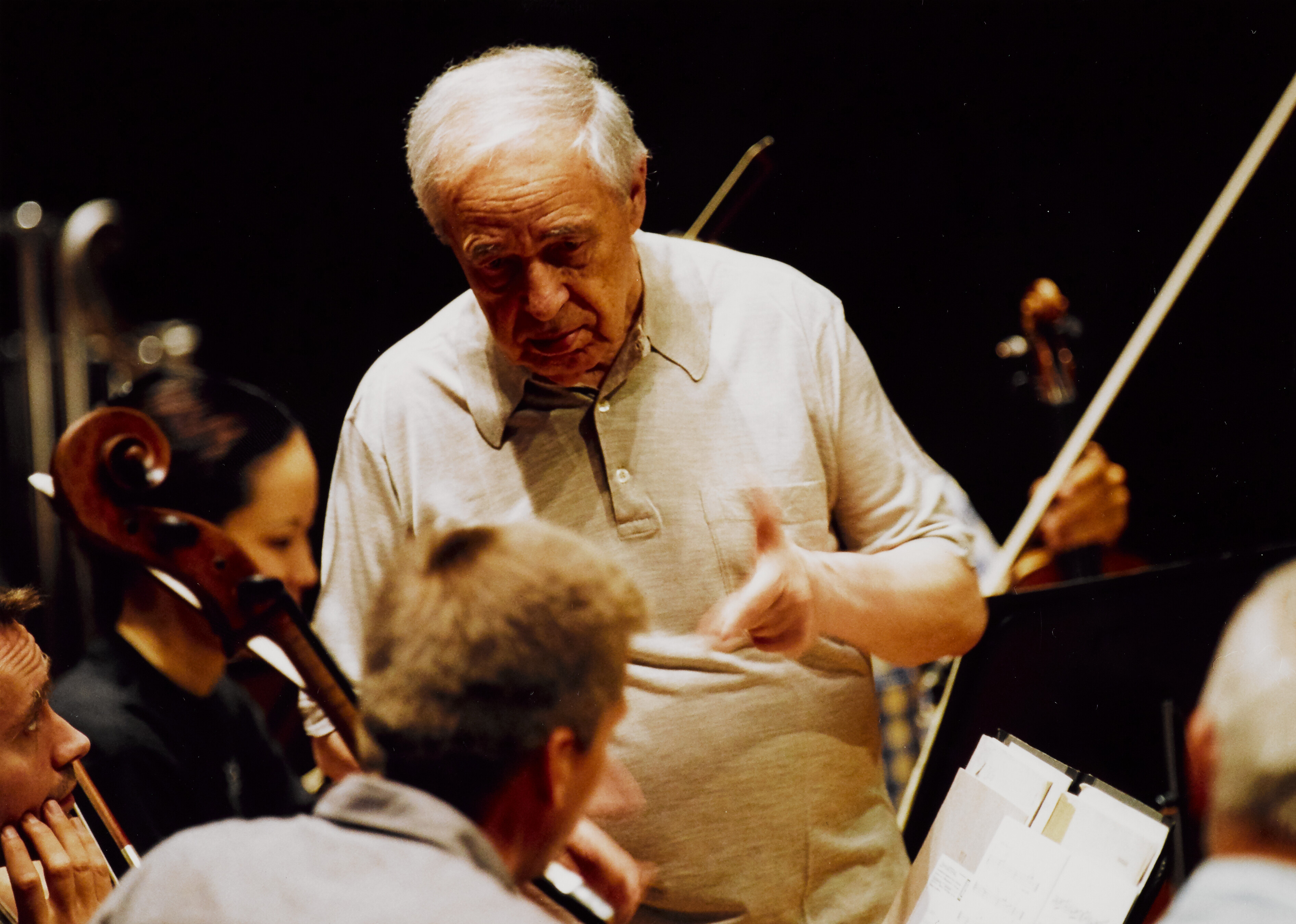 Pierre Boulez in einem hellen Polohemd dirigiert die Musiker während einer Probe, wobei er mit der Hand gestikuliert, während mehrere Orchestermitglieder zuhören und auf die Notenblätter schauen.