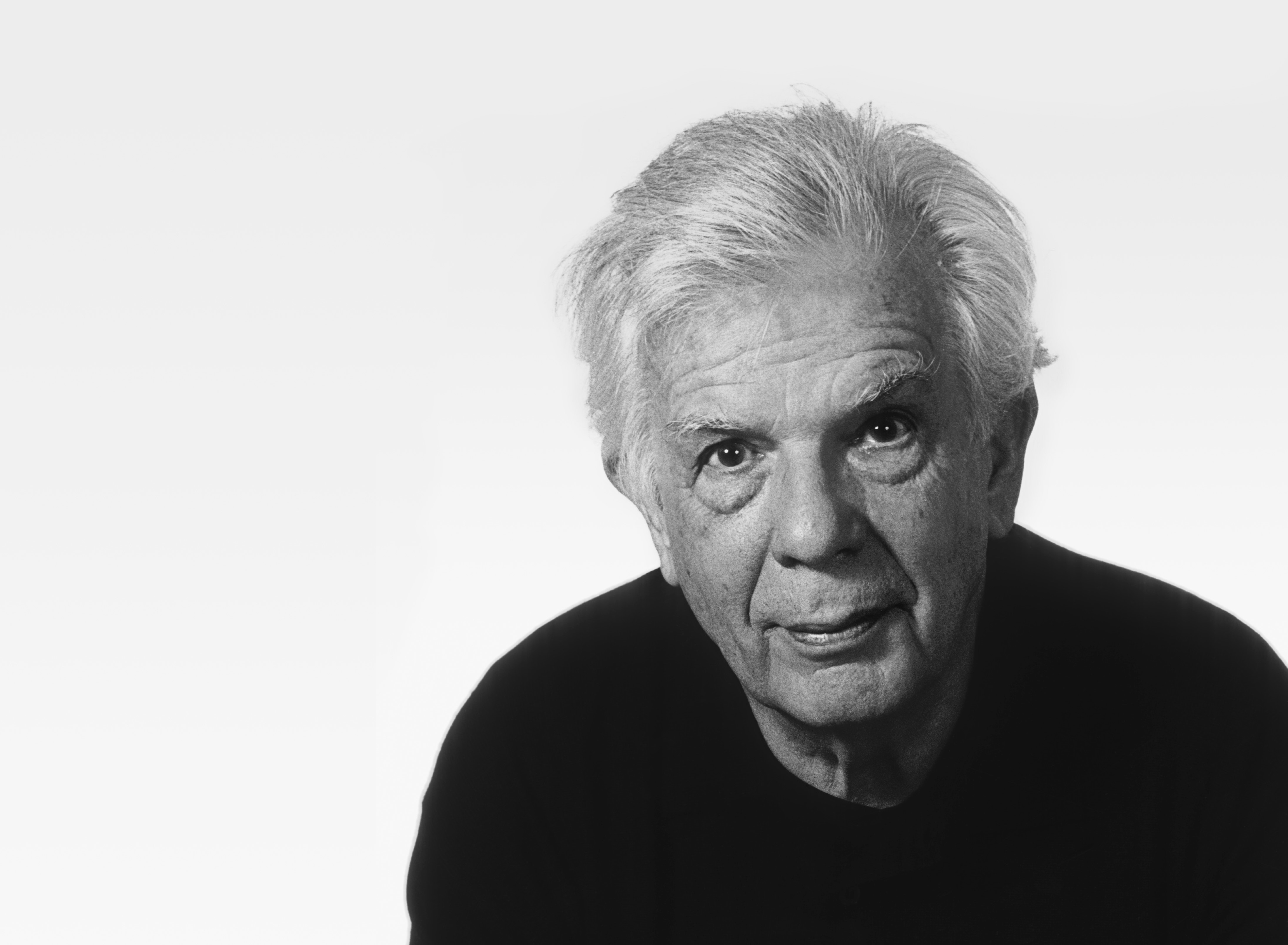 Black and white portrait of an older man with white hair, wearing a dark shirt, looking directly at the camera with a neutral expression against a plain light background.