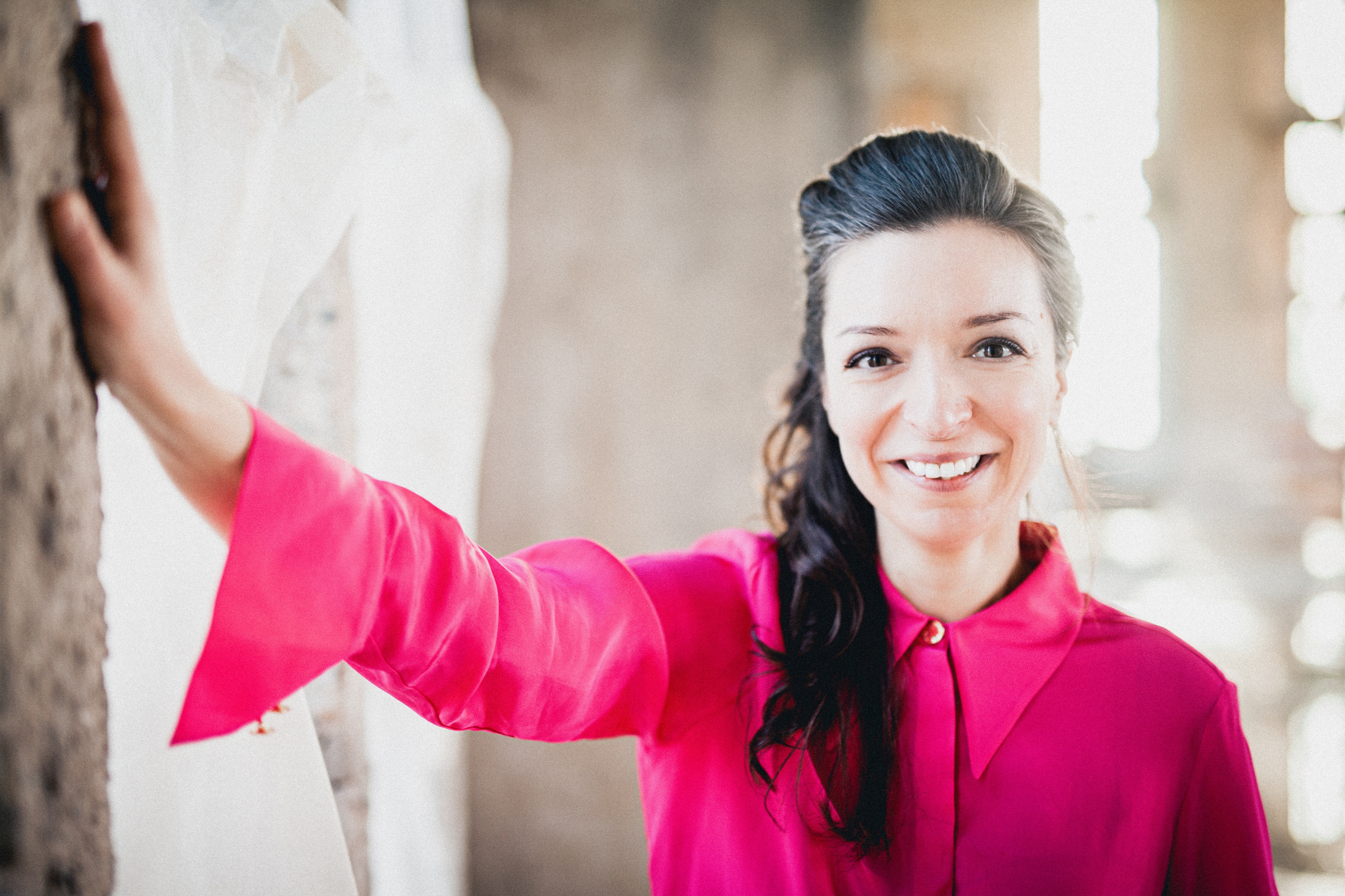 Yulianna Avdeeva  in einer leuchtend rosa Bluse steht im Innenbereich und stützt ihre Hand an die Wand. Der Hintergrund ist leicht verschwommen, was ihren fröhlichen Gesichtsausdruck und die leuchtenden Farben der Kleidung hervorhebt.