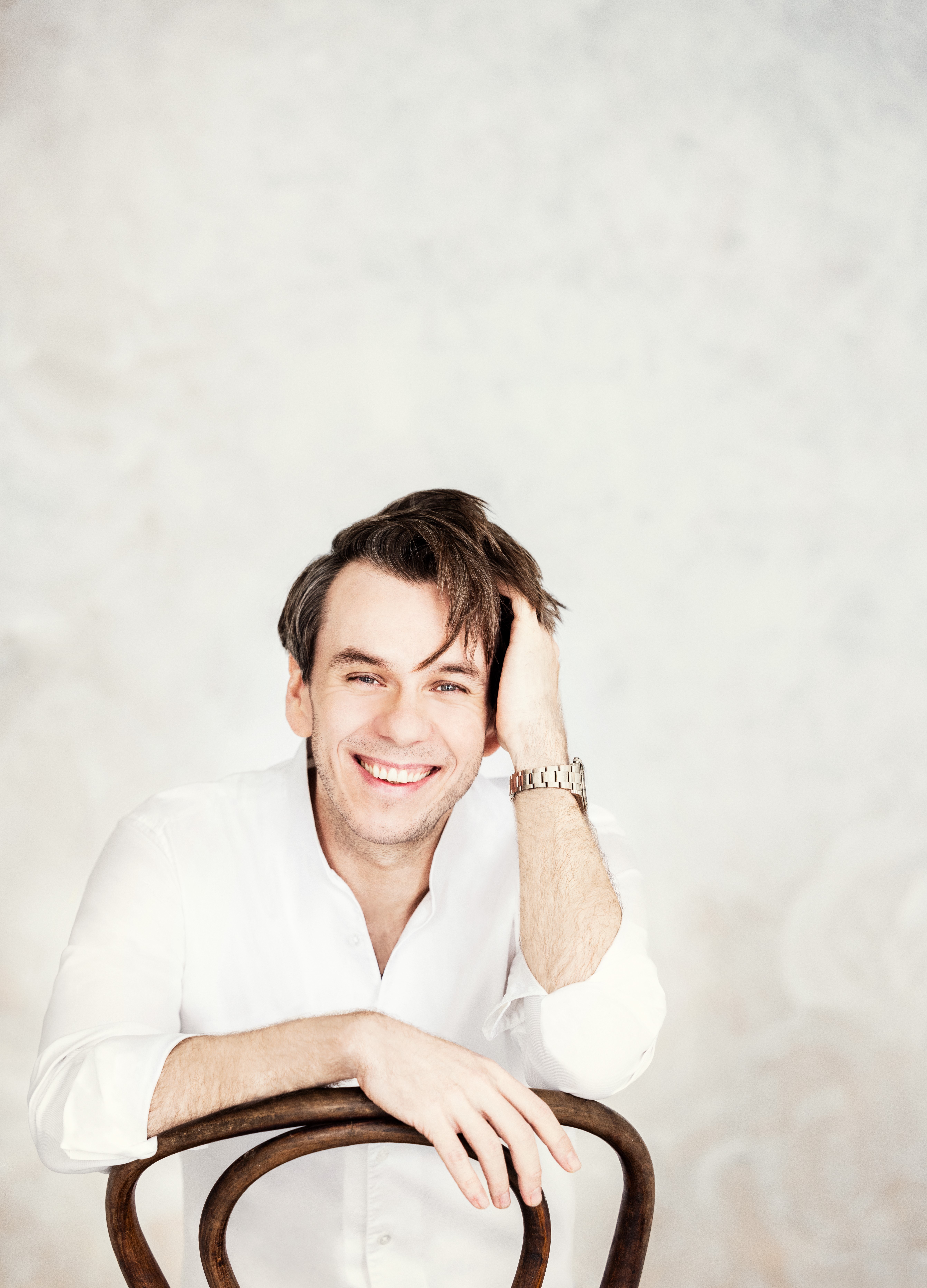 Benjamin Bernheim sits smiling on a chair, wearing a white shirt and a watch, with his hand resting on his head, set against a bright, textured background.