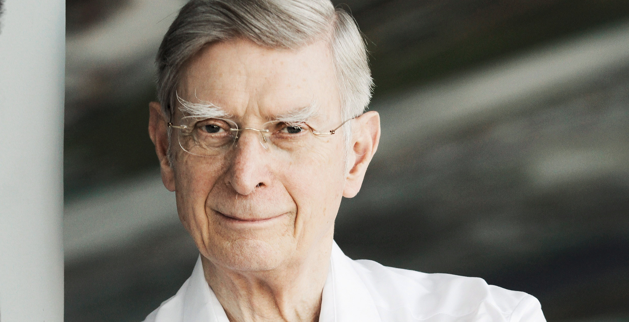 Herbert Blomstedt mit grauem Haar und Brille, trägt ein weißes Hemd und lächelt sanft, während er in die Kamera blickt. Der Hintergrund ist unscharf.