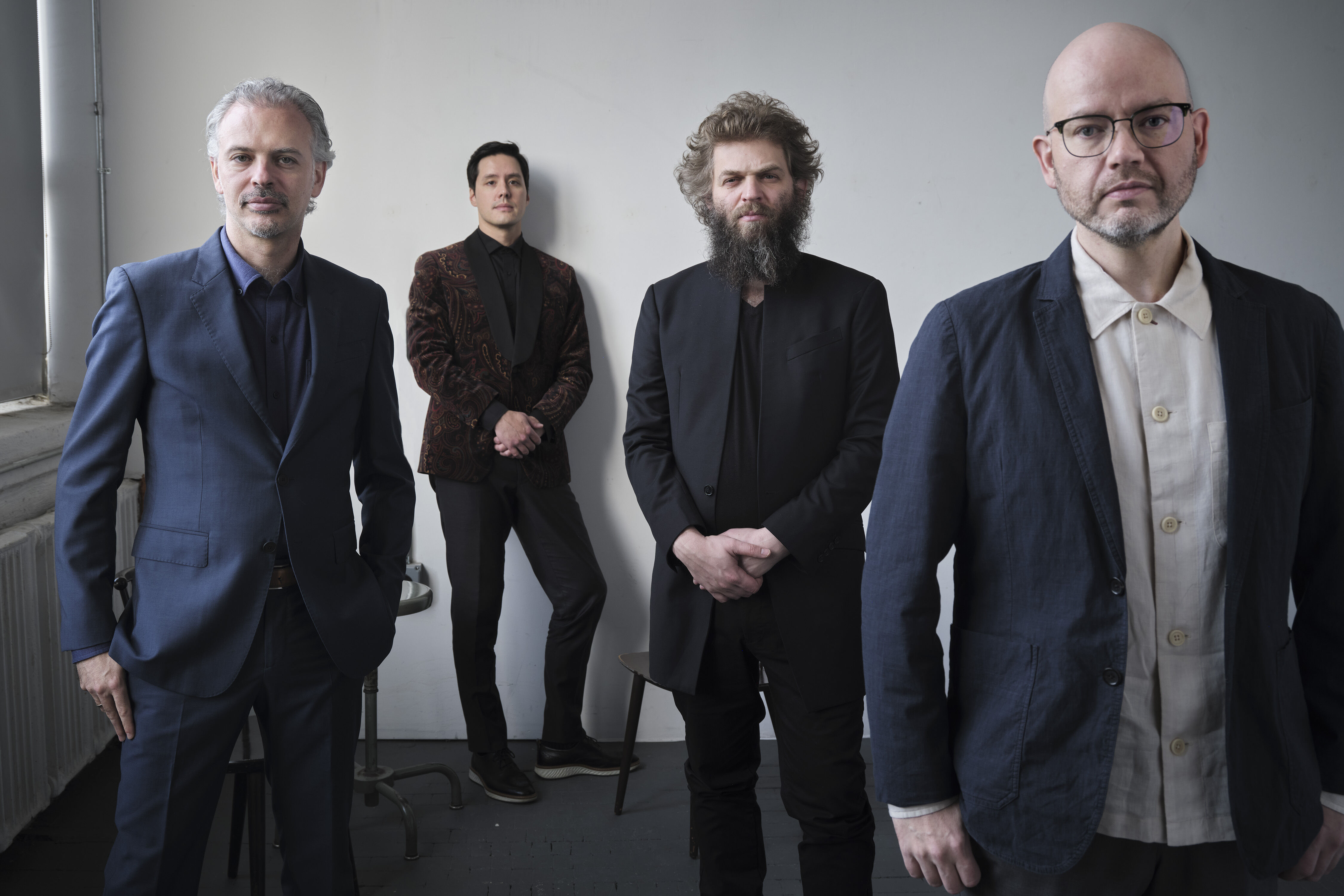 Vier Männer in Anzügen stehen verteilt in einem Raum vor einer weißen Wand. Sie schauen in die Kamera.