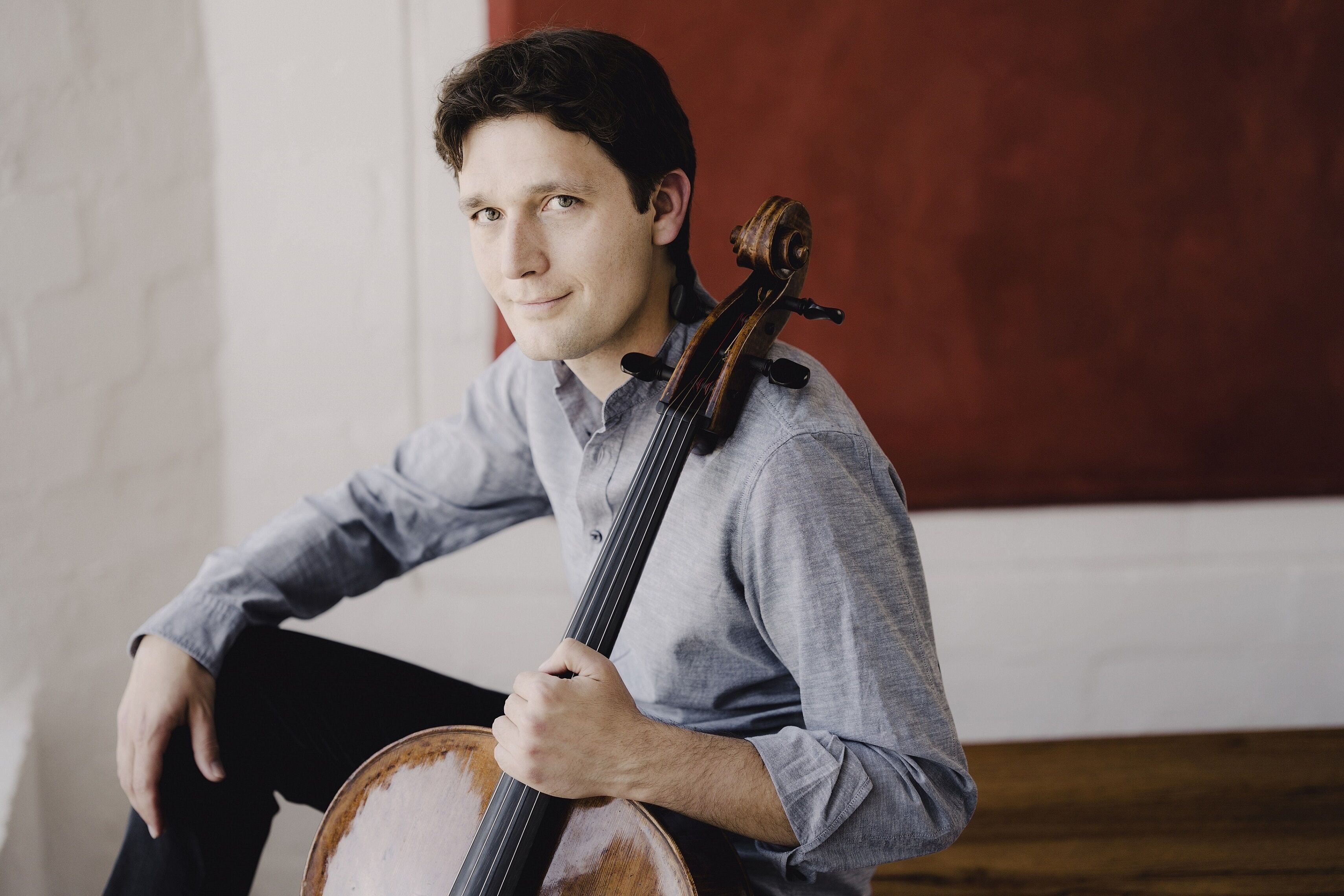 Junger Mann mit dunklem Haar in grauem Hemd, hält ein Cello und blickt freundlich in die Kamera. Er sitzt entspannt vor einer weißen Wand mit rotem Hintergrundelement.