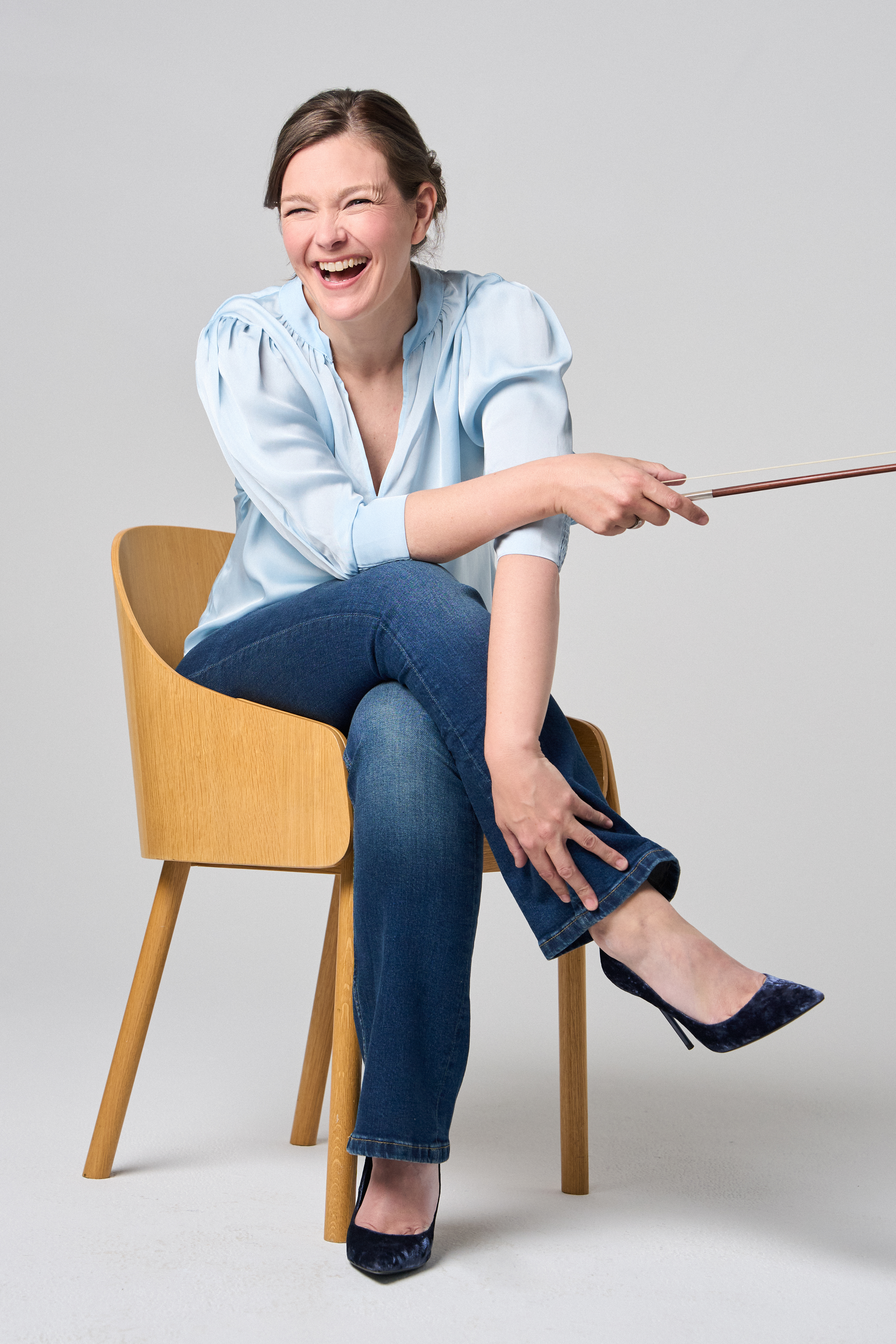 Janine Jansen is sitting on a chair, a violin in her hands. She is wearing jeans and a light blue blouse and looks into the camera with a smile.