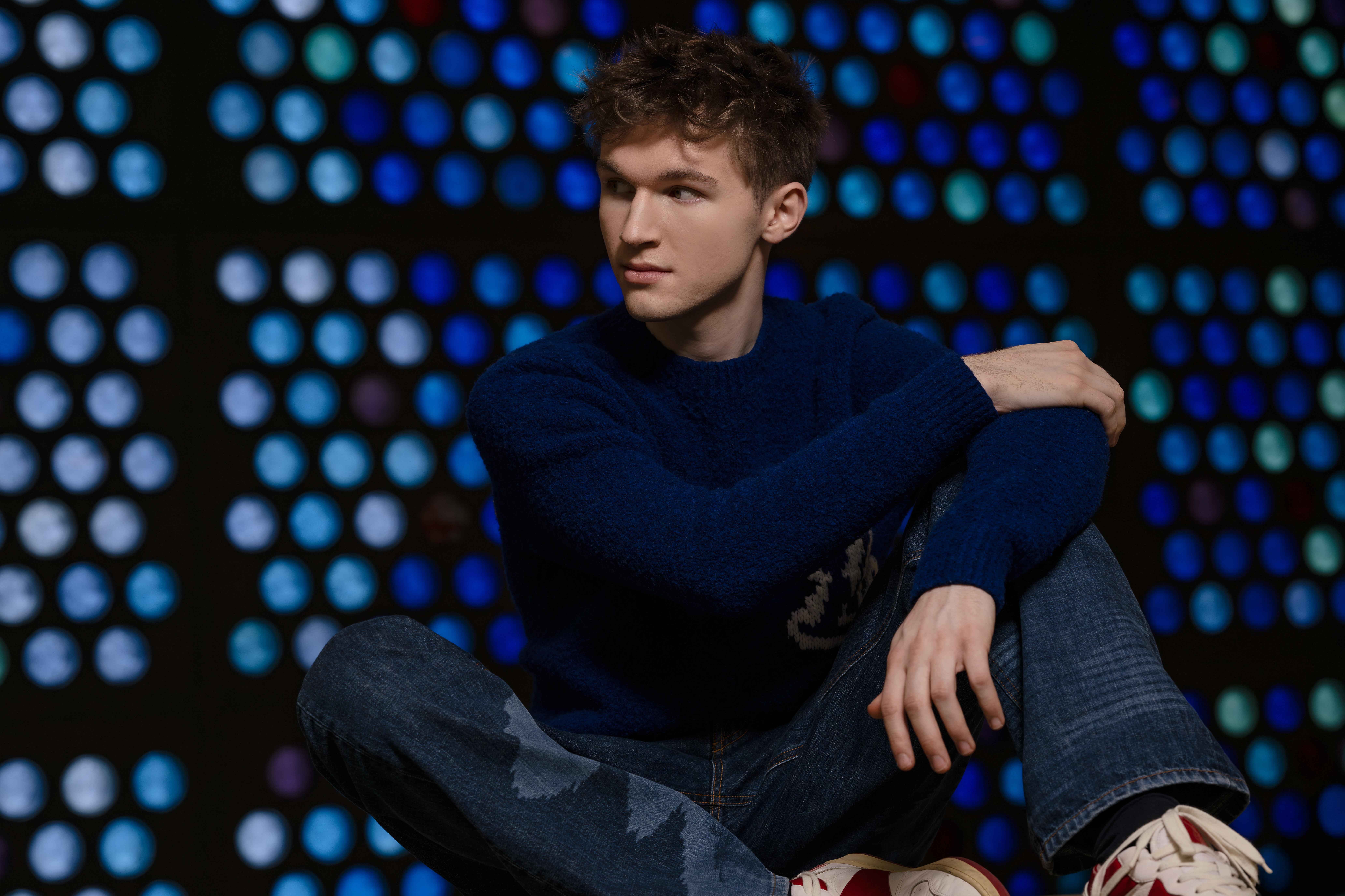 Portrait of the young man Jan Liebermann, sitting in a blue jumper and jeans in front of a background with blue, round glass windows. He is looking to the side. 