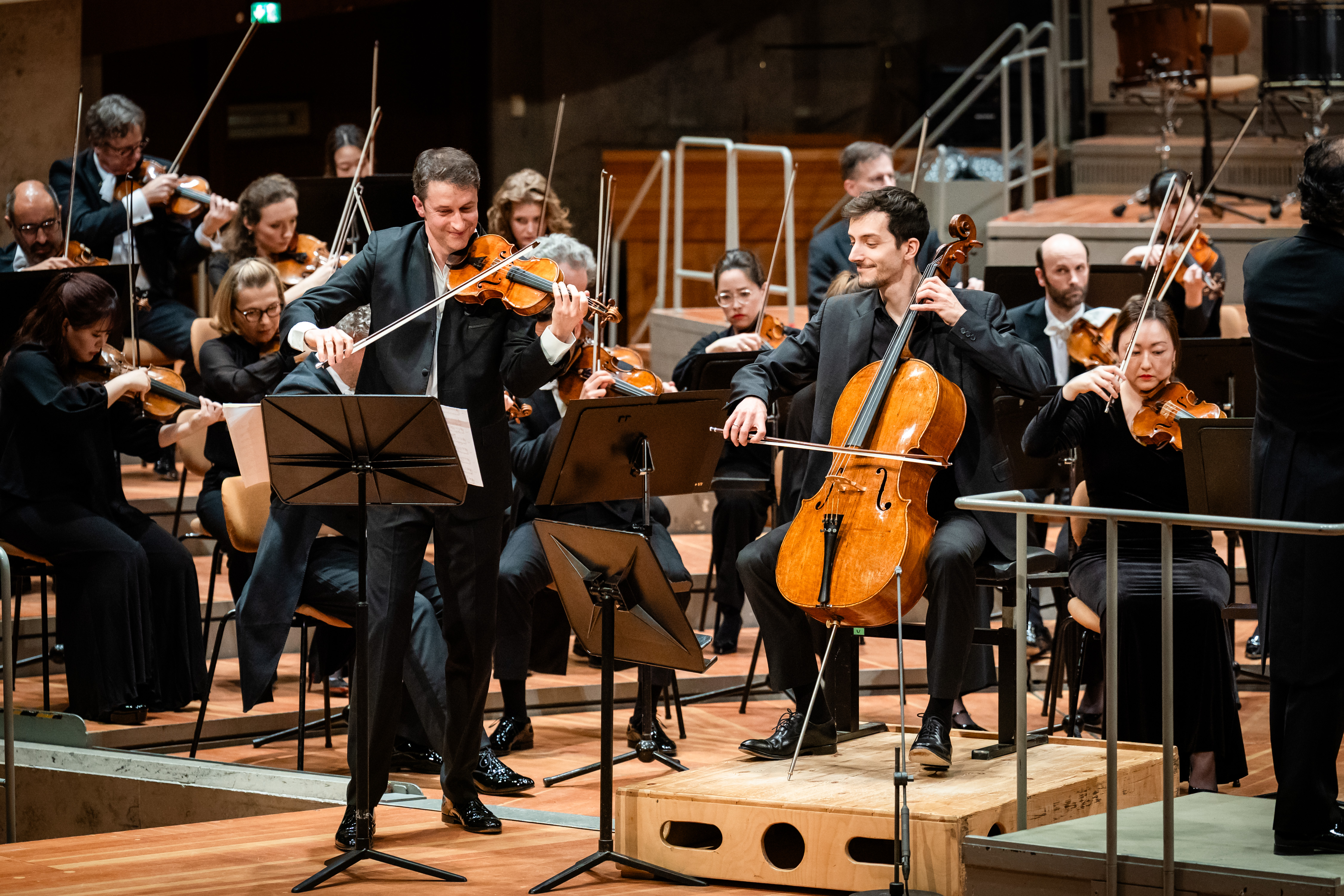 An orchestra performs on stage. Noah Bendix-Balgley und Bruno Delepelaire play at the front, surrounded by other musicians with string instruments. Music stands and sheet music are visible.