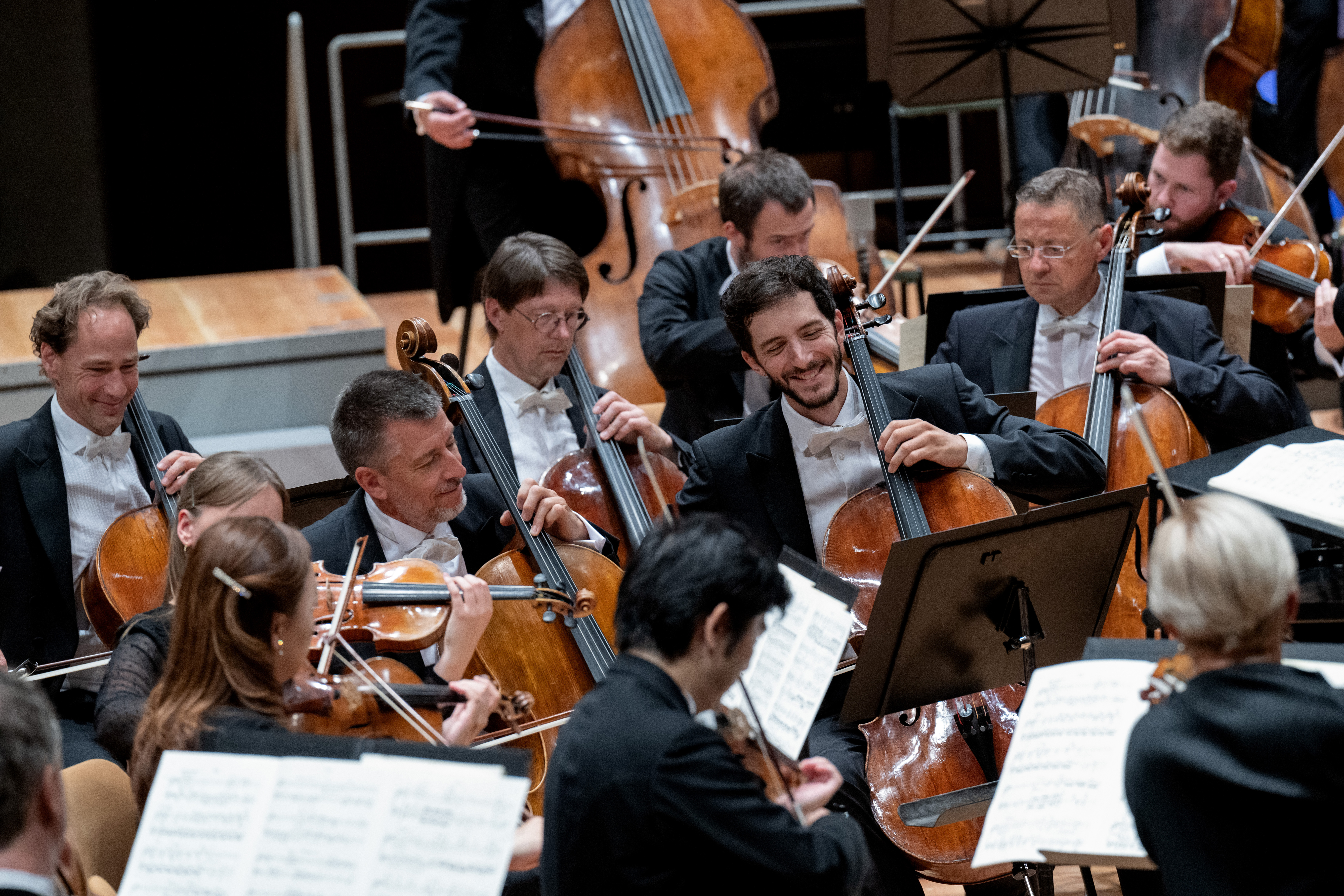 Mehrere Musikerinnen in formeller Kleidung spielen Celli und Geigen in einem Orchester. Sie sitzen eng beieinander, lesen Noten und einige lächeln, während sie auf der Bühne spielen.