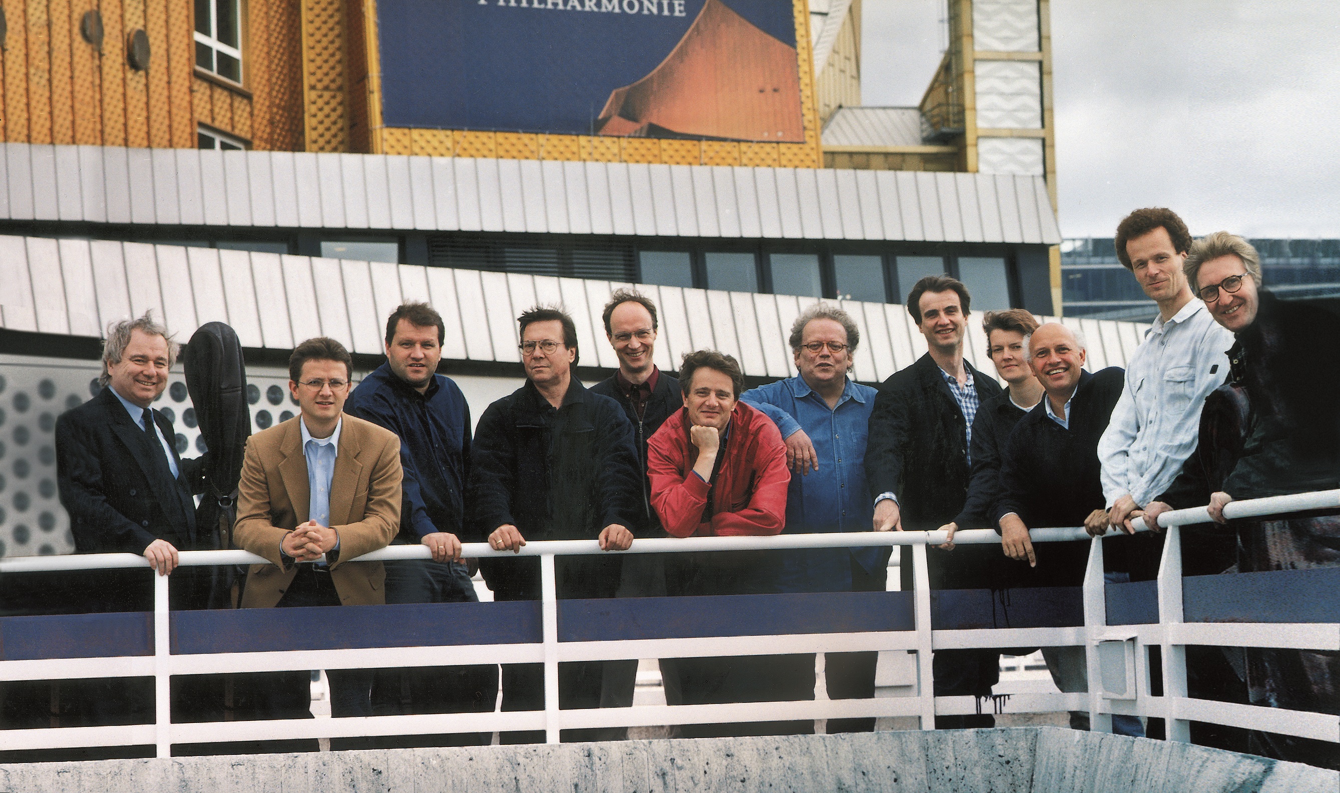 Zwölf Männer stehen, an ein weißes Geländer gelehnt, lächelnd im Freien vor einem modernen Gebäude mit gelben und weißen Tafeln.