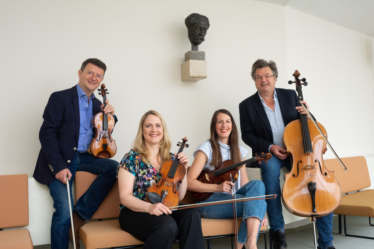 Das Bild zeigt vier lächelnde Musiker mit ihren Streichinstrumenten: zwei Geigen, einer Bratsche und einem Cello. Sie sitzen oder stehen entspannt in einem hellen Raum, an der Wand hinter ihnen hängt eine Büste.