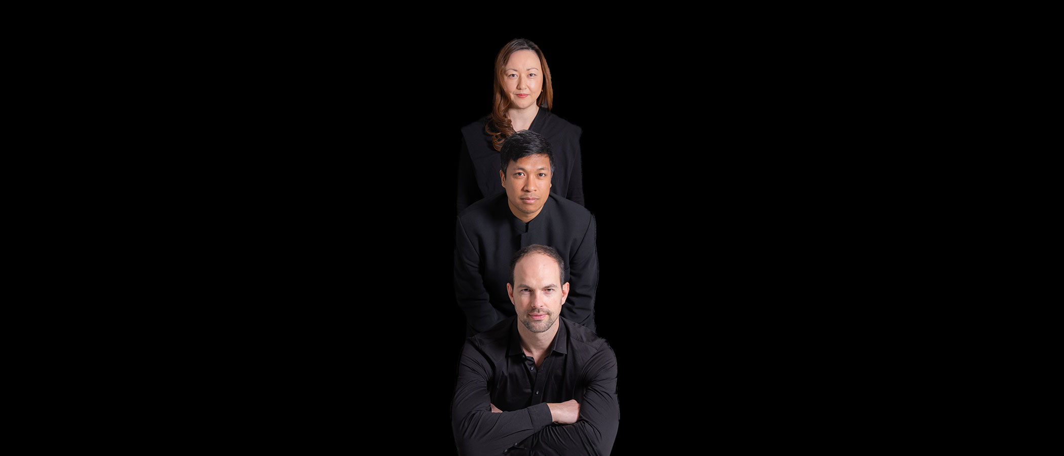 Three people in black clothing are posed in a vertical line against a black background, with a man in front, another man behind him, and a woman at the top, all facing forward.