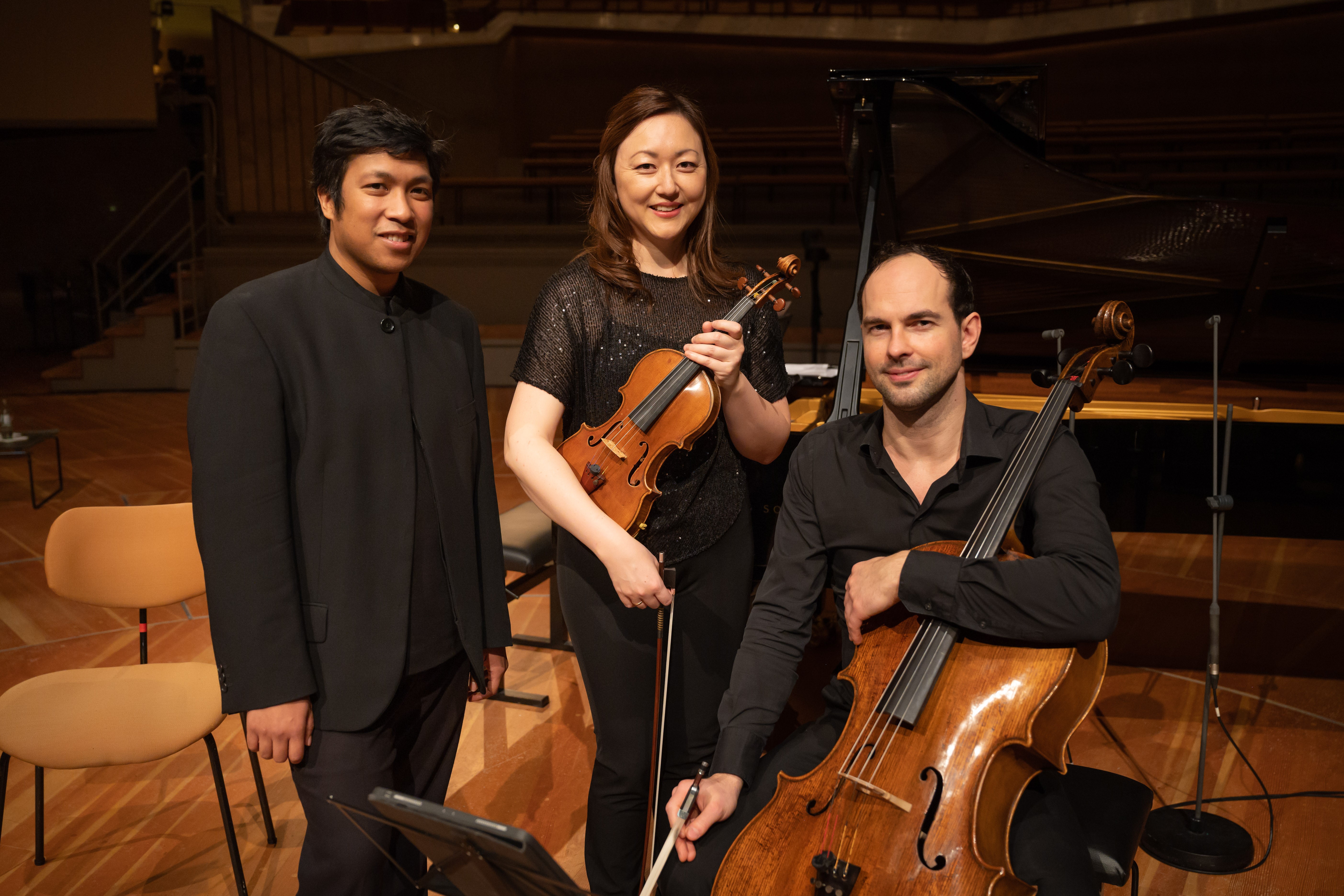 Drei Musiker posieren auf einer Holzbühne. Einer hält eine Geige, ein anderer ein Cello, der dritte steht daneben. Im Hintergrund sind ein Klavier und leere Sitze zu sehen, die eine entspannte Konzertatmosphäre schaffen.
