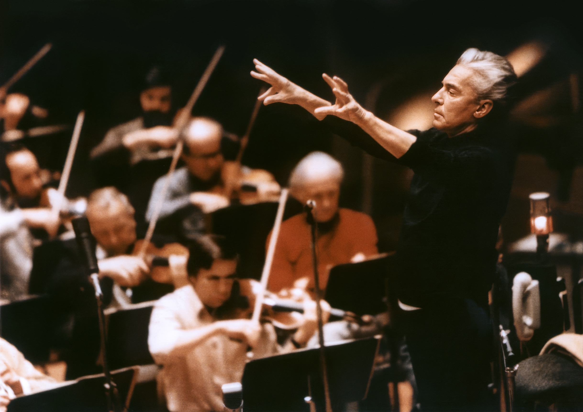 Herbert von Karajan steht mit erhobenen Armen und leitet leidenschaftlich die Berliner Philharmoniker, die in einem schwach beleuchteten Konzertsaal Streichinstrumente spielen.