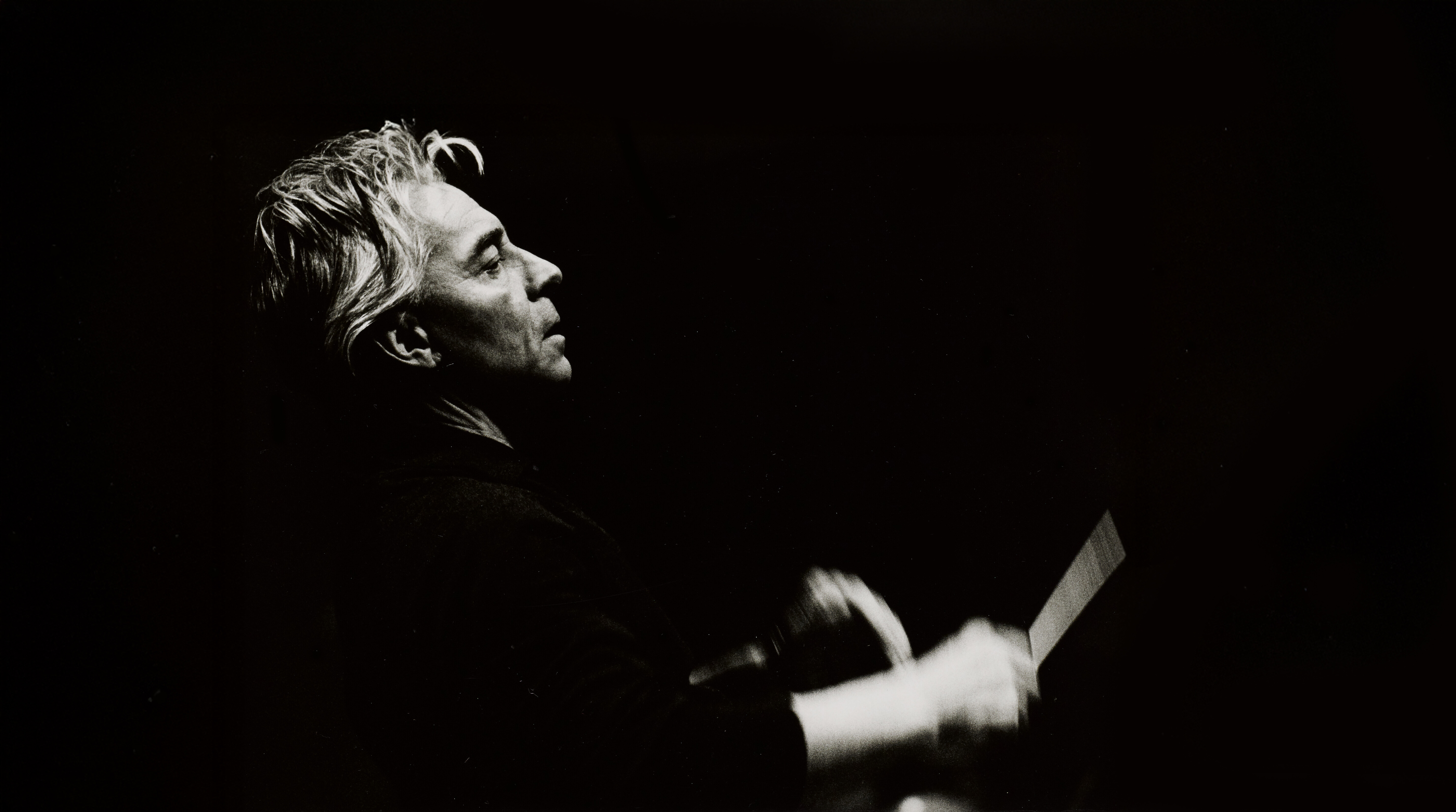 Black-and-white photo of a person with light hair, seen in profile, conducting with an expressive gesture, set against a dark background.