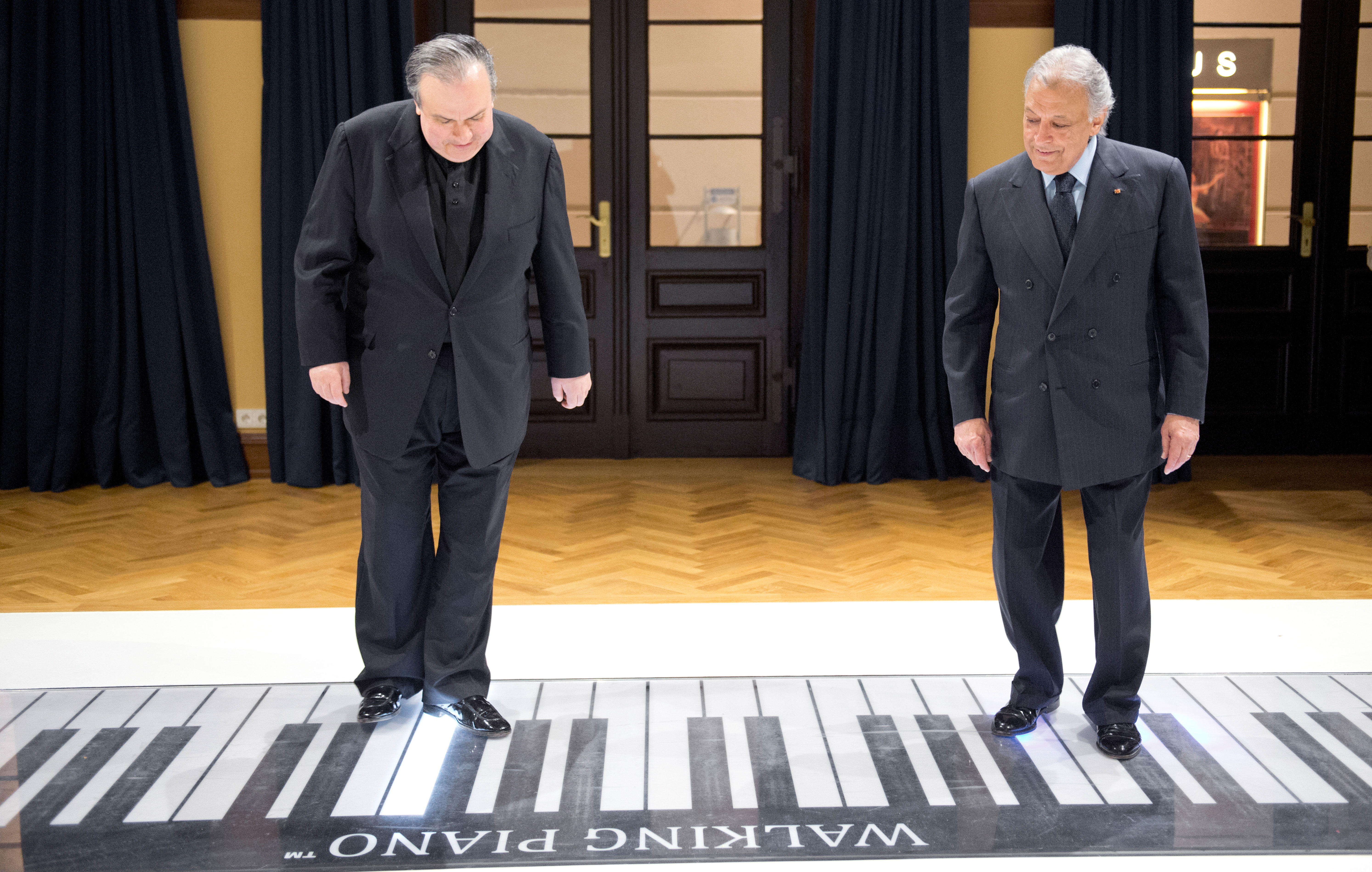 Zwei Männer in Anzügen stehen an einer großen Klaviertastatur und schauen auf die Tasten hinunter. Die Szene scheint in einem Innenraum mit Holzfußboden, blauen Vorhängen und dunklen Holztüren hinter ihnen zu spielen.