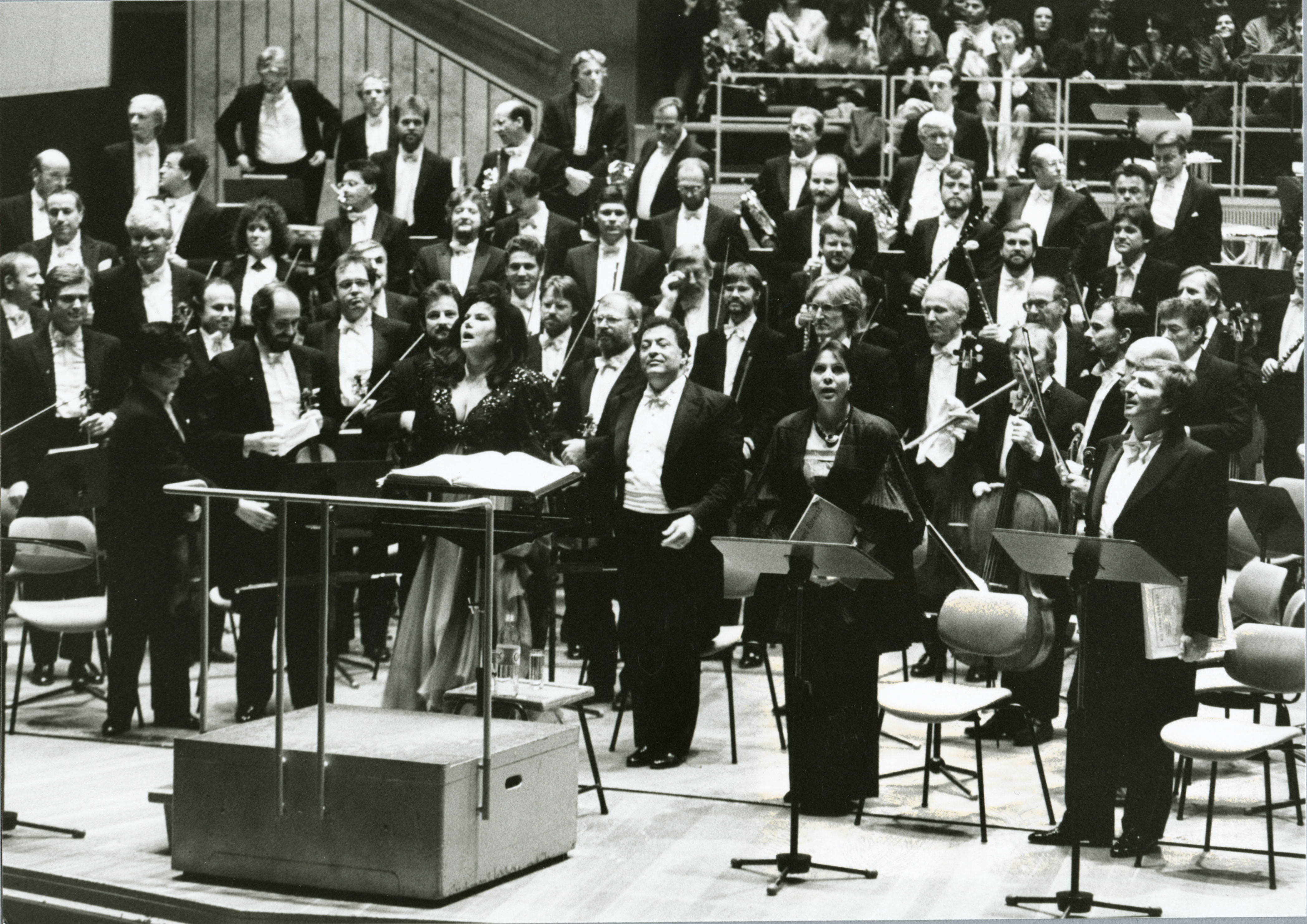 Ein großes Orchester und mehrere Gesangssolisten stehen in förmlicher Kleidung auf der Bühne, sind dem Publikum zugewandt und scheinen den Applaus in einem Konzertsaal zu quittieren, während im Hintergrund Zuschauer zu sehen sind.