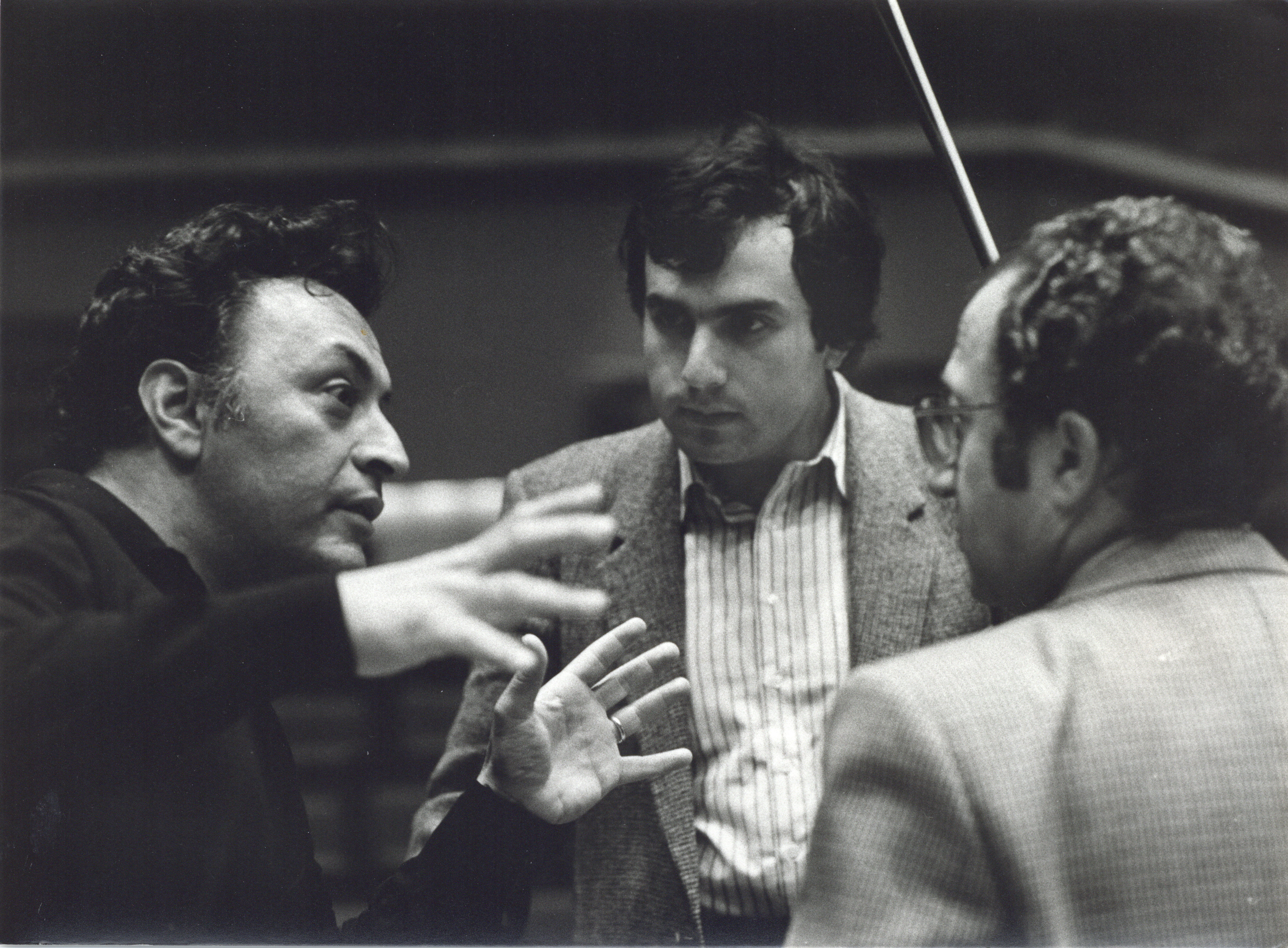 Drei Männer stehen in einem Haus und führen eine lebhafte Diskussion. Ein Mann gestikuliert ausdrucksstark mit seinen Händen, während die beiden anderen aufmerksam zuhören. Alle sind in Jacken gekleidet, und über ihnen ist ein Dirigentenstab zu sehen.