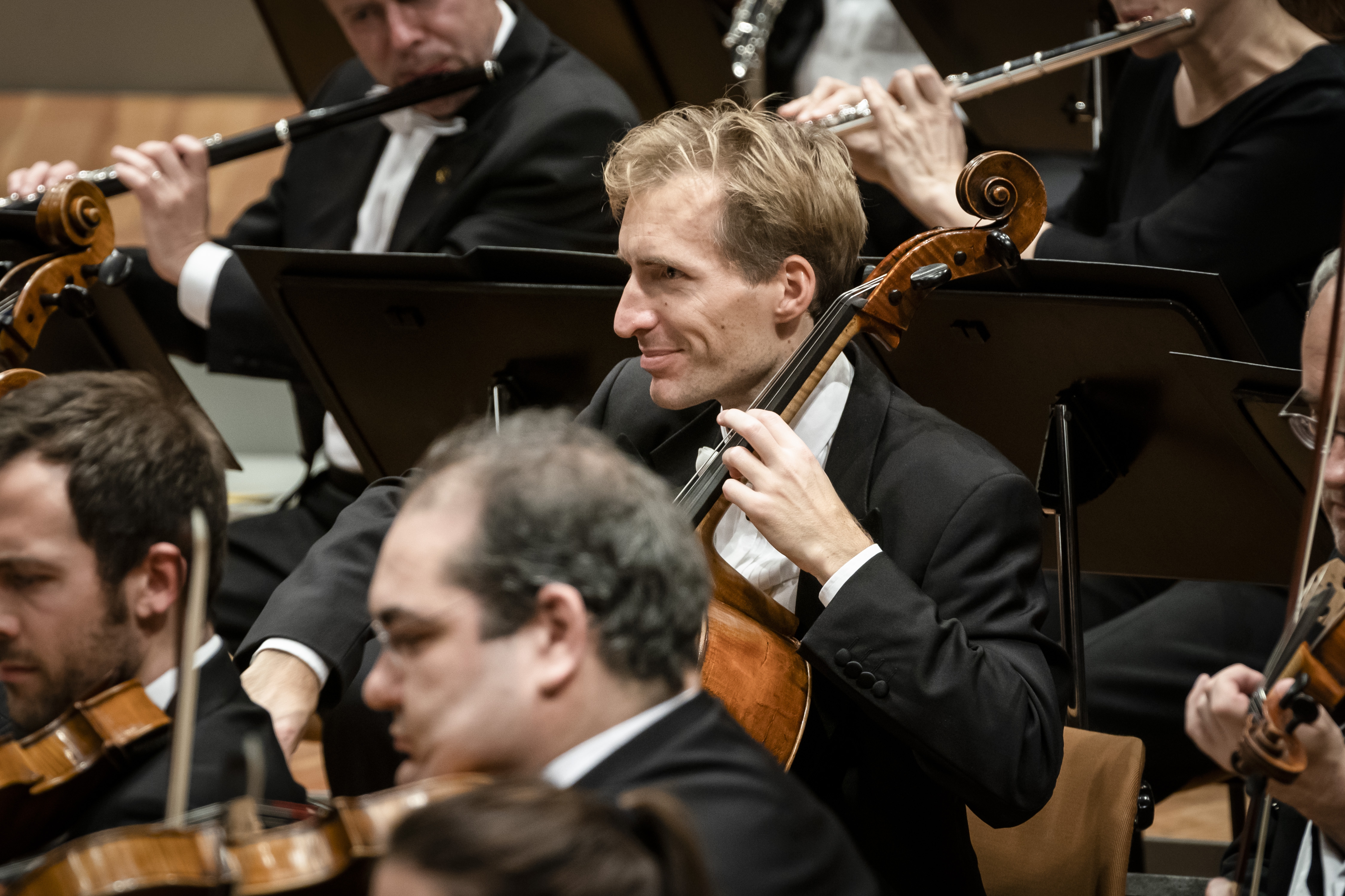 Ein Mann in einem schwarzen Frack spielt Cello in einem Orchester, umgeben von anderen Musikern, die auf der Bühne Streich- und Blasinstrumente spielen.