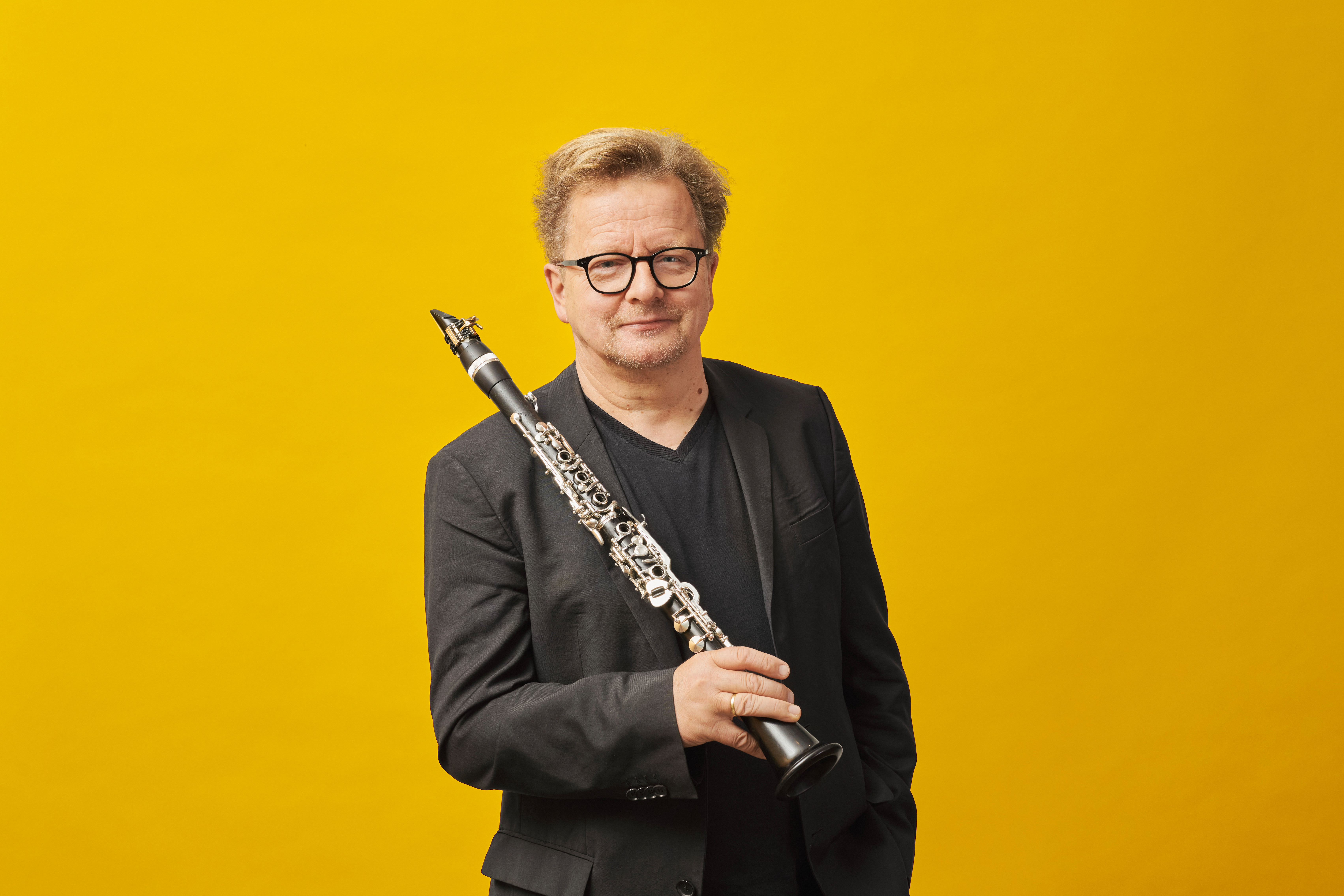 A portrait photo of Wenzel Fuchs, standind in front of a yellow background with his clarinet in his hands.
