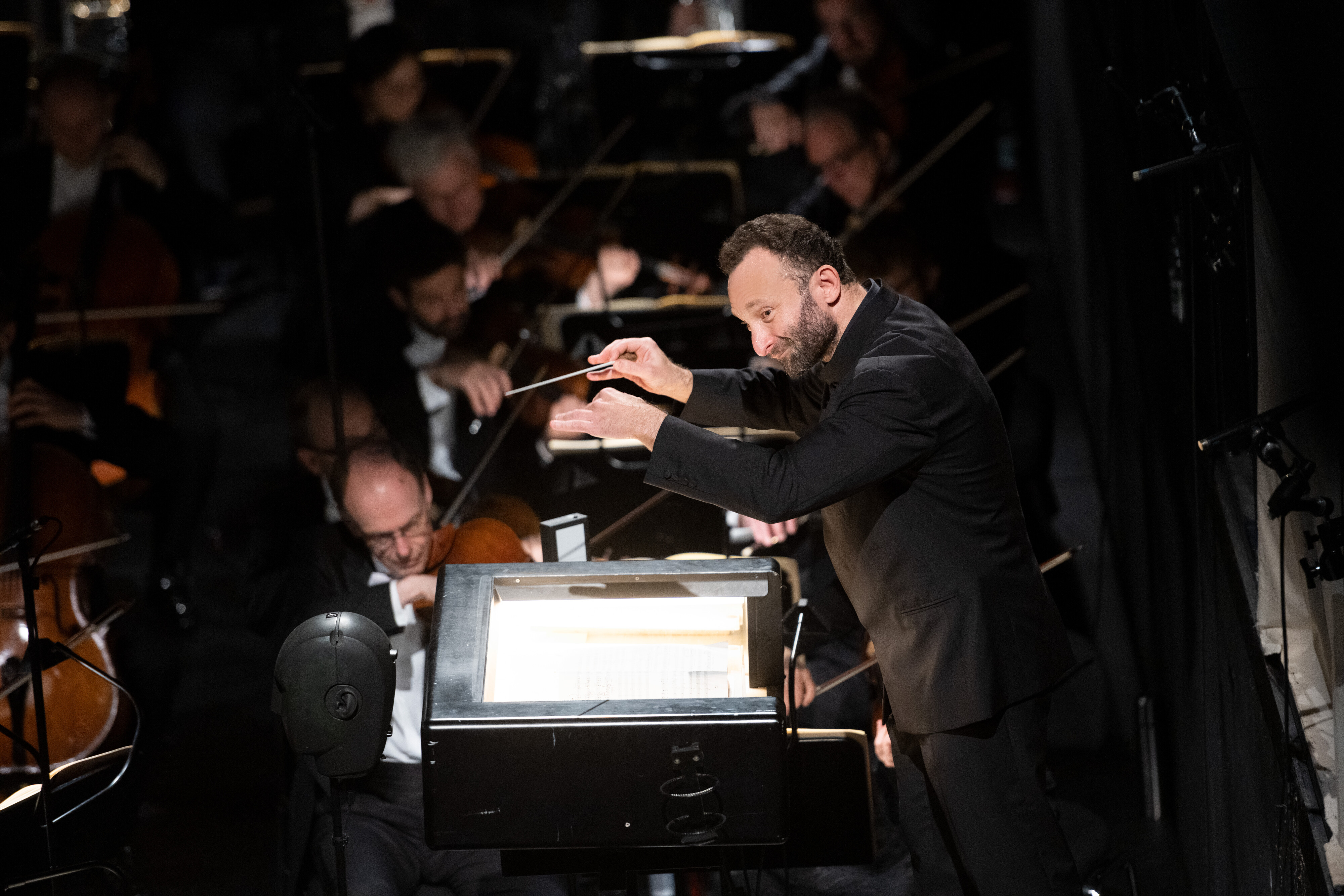 Kirill Petrenko in a black suit energetically leads an orchestra, holding a baton, with musicians playing string instruments in the background and an open score on the stand in front of him.
