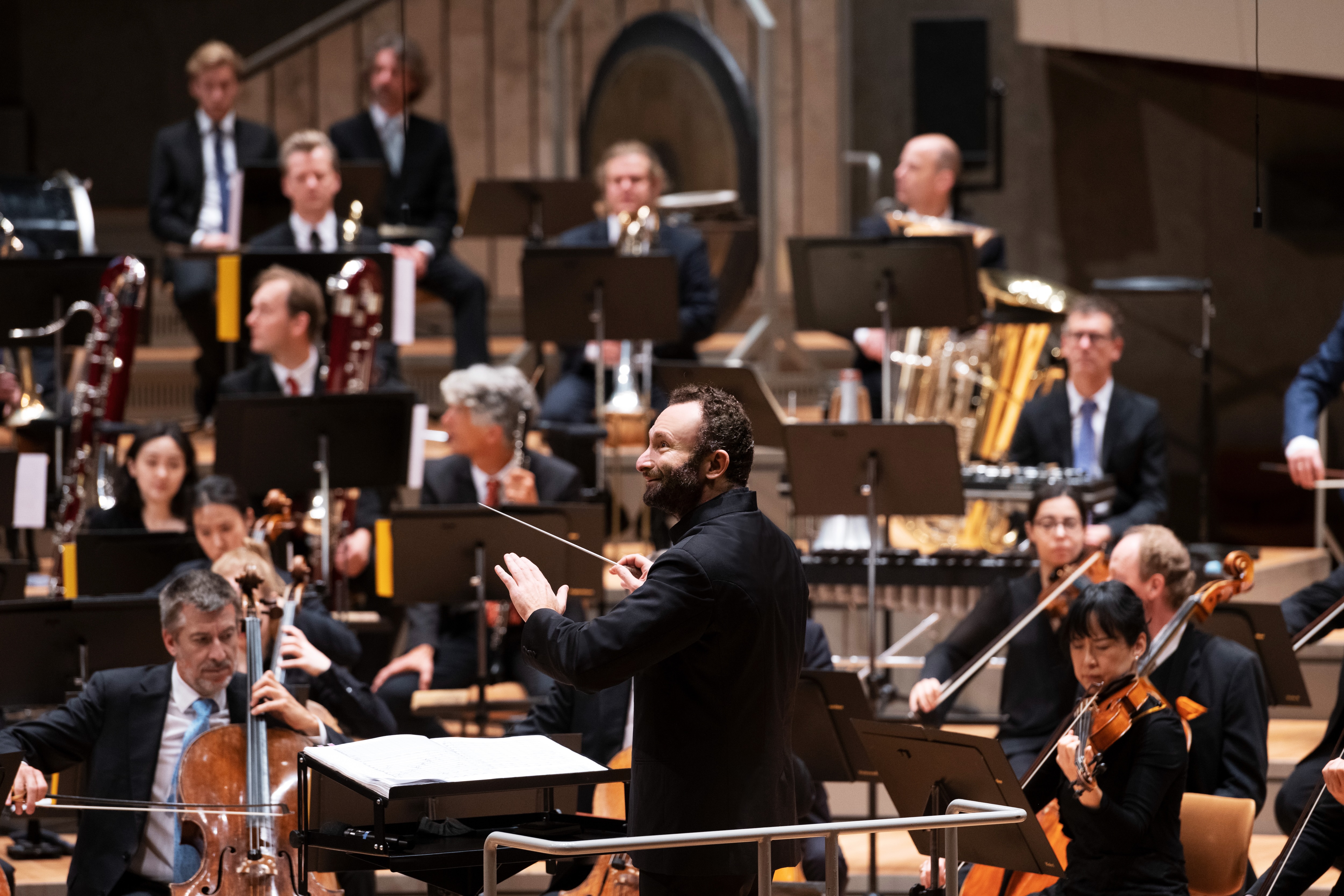 Ein Dirigent leitet ein Orchester während einer Aufführung; die Musiker spielen verschiedene Instrumente, darunter Streicher und Blechbläser, während sie die Noten auf Ständern lesen.