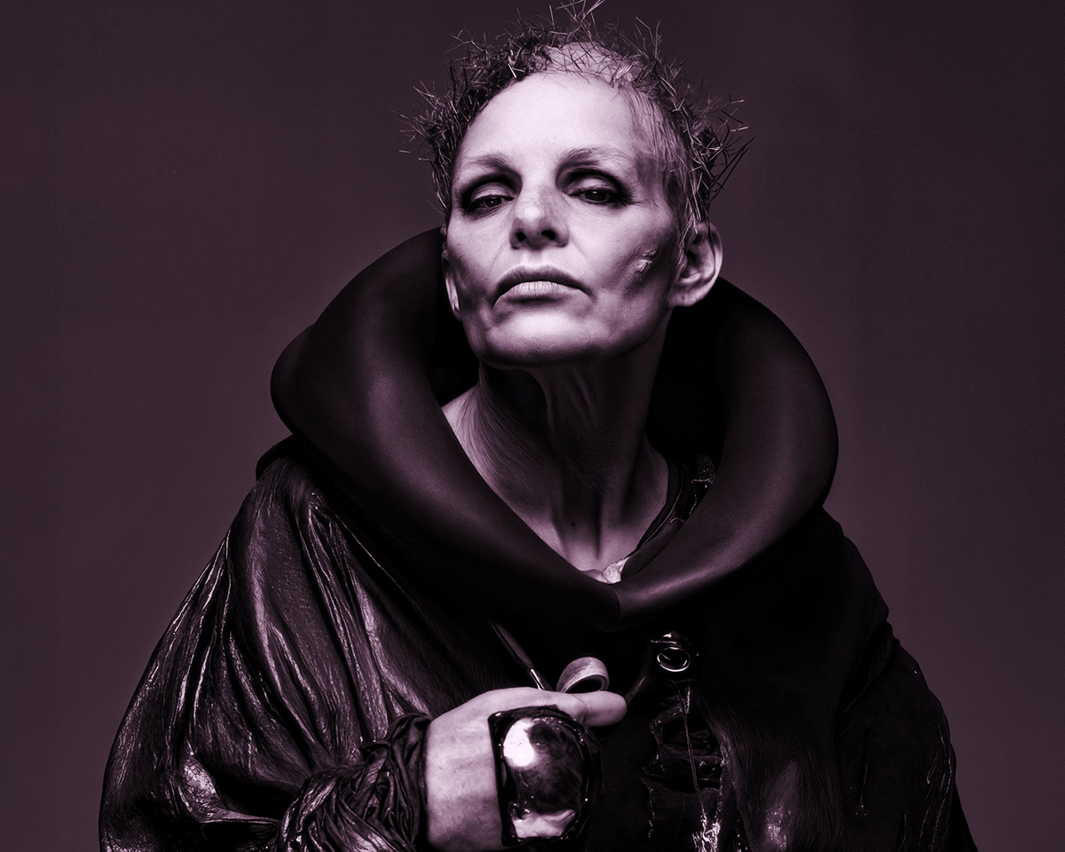 Androgyne Person mit scharfen Wangenknochen, strukturiertem kurzem Haar und einem dramatischen dunklen Outfit mit hohem Kragen starrt selbstbewusst in die Kamera. Das Bild hat einen monochromen violetten Farbton.
