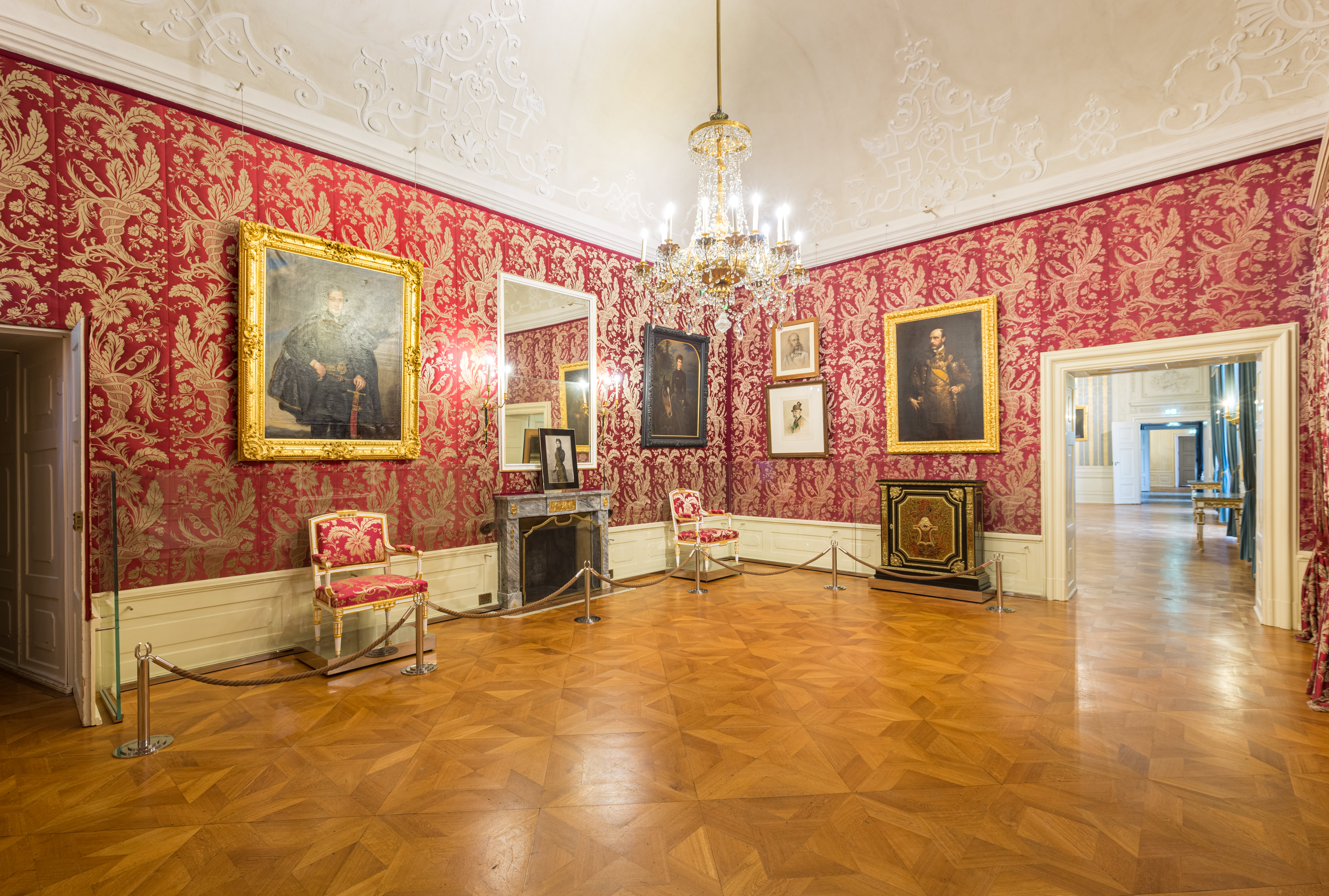Ein prunkvoller Raum mit roter und goldener Damasttapete, verzierten Stühlen, einem Kronleuchter, gerahmten Porträts, Parkettboden und vergoldetem Dekor, betrachtet vom Eingang eines historischen Palastes oder Herrenhauses.