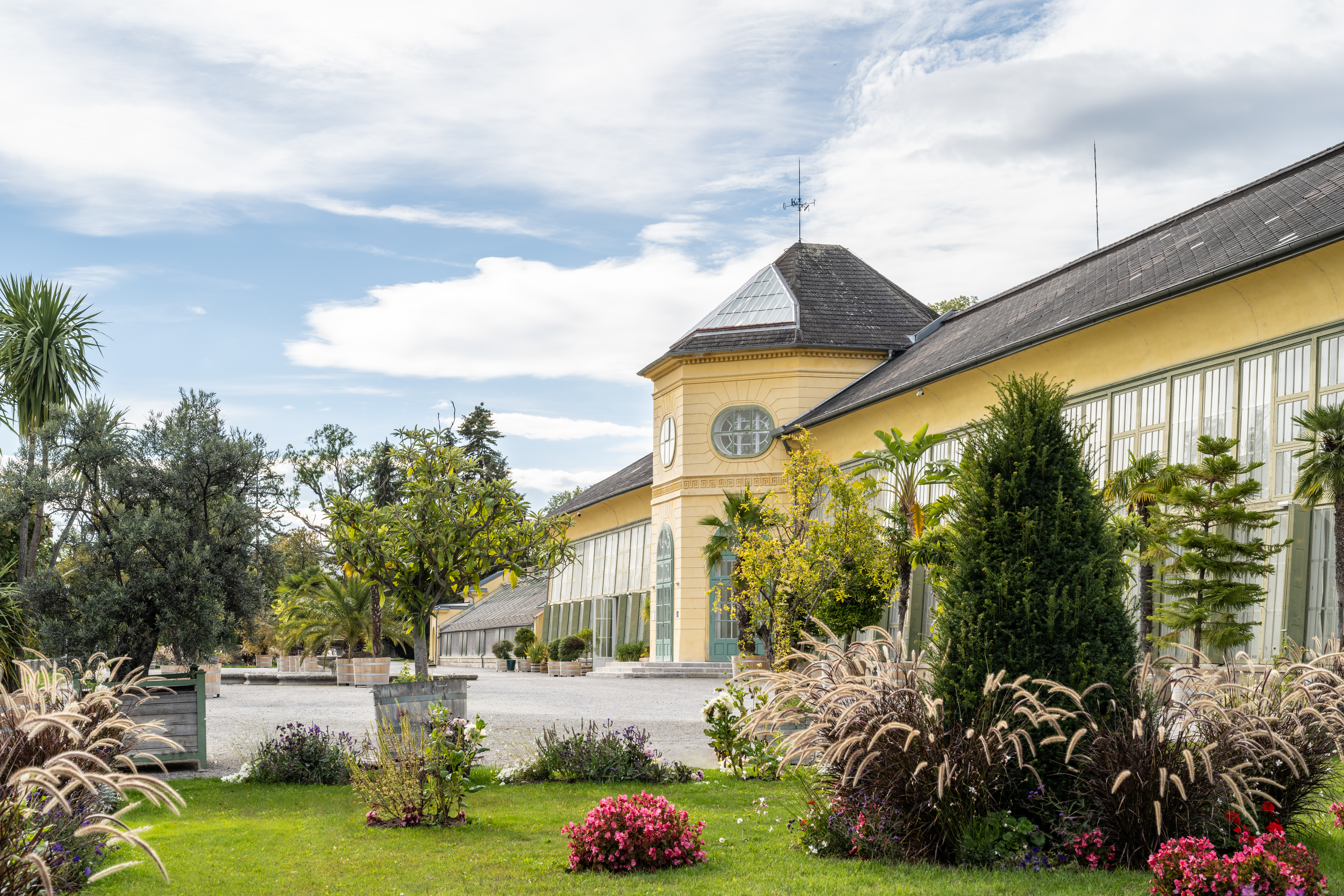 Ein großes gelbes Gewächshaus mit Glasfenstern und einem Uhrenturm steht inmitten grüner Bäume, blühender Pflanzen und gepflegter Gärten unter einem teilweise bewölkten Himmel.