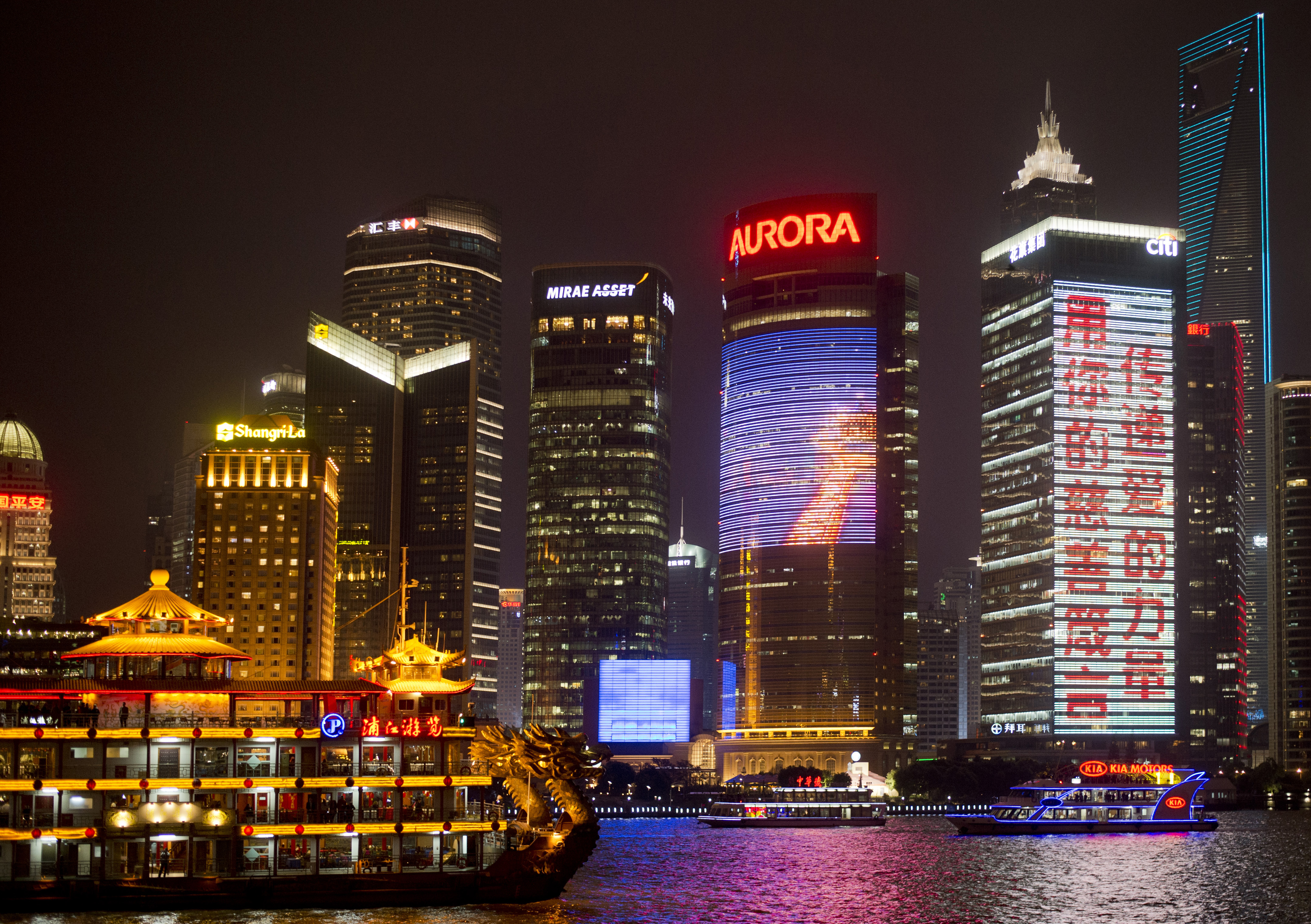 Bunt beleuchtete Boote treiben nachts auf einem Fluss vor der Skyline Shanghais mit ihren hohen Wolkenkratzern, die von hellen Schildern und elektronischen Werbetafeln beleuchtet werden.