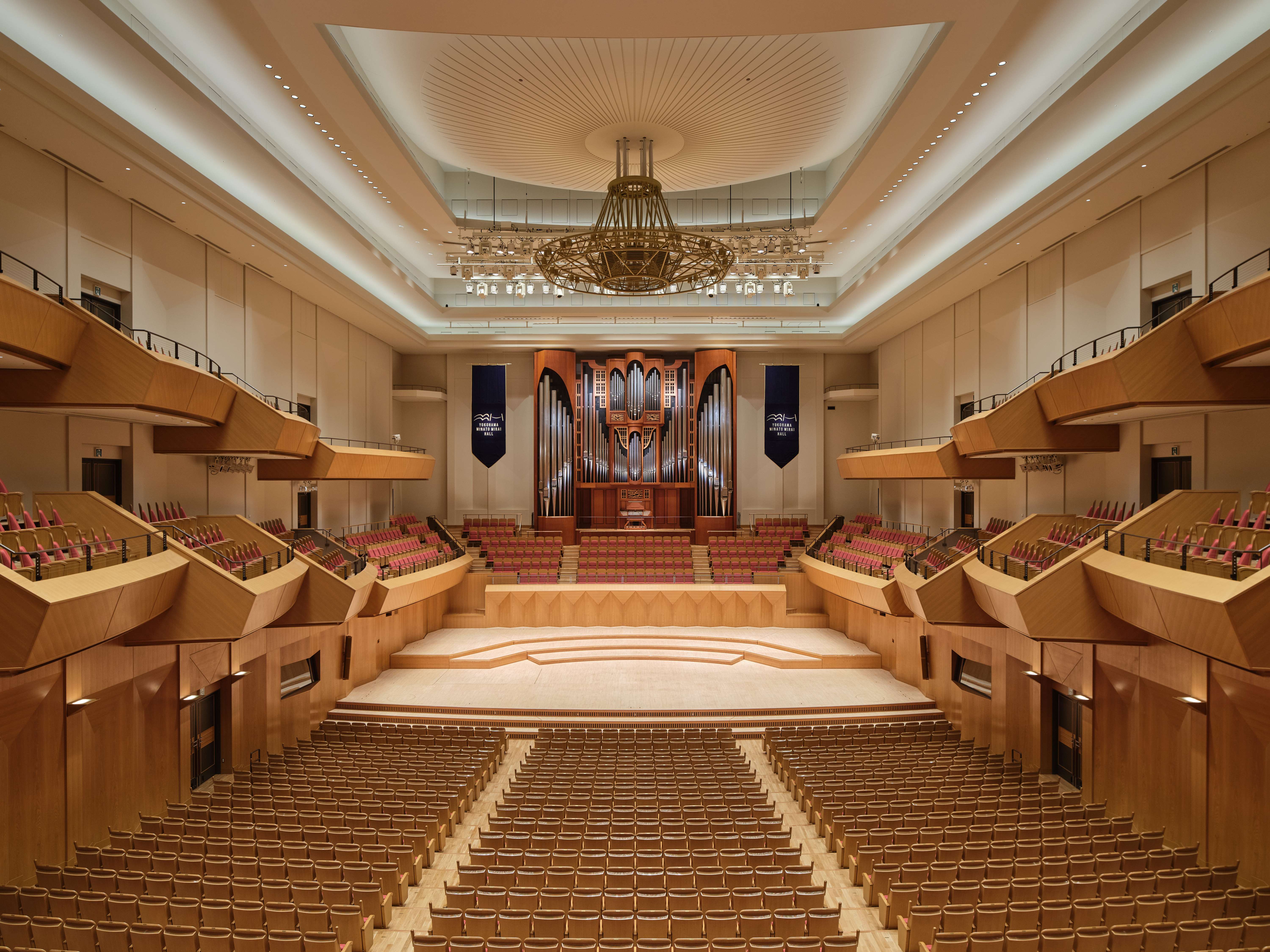Ein geräumiger, moderner Konzertsaal mit Sitzreihen, einer großen Pfeifenorgel im hinteren Teil der Bühne, Holzvertäfelung und einem dekorativen Kronleuchter in der Mitte unter einer hohen, gewölbten Decke.