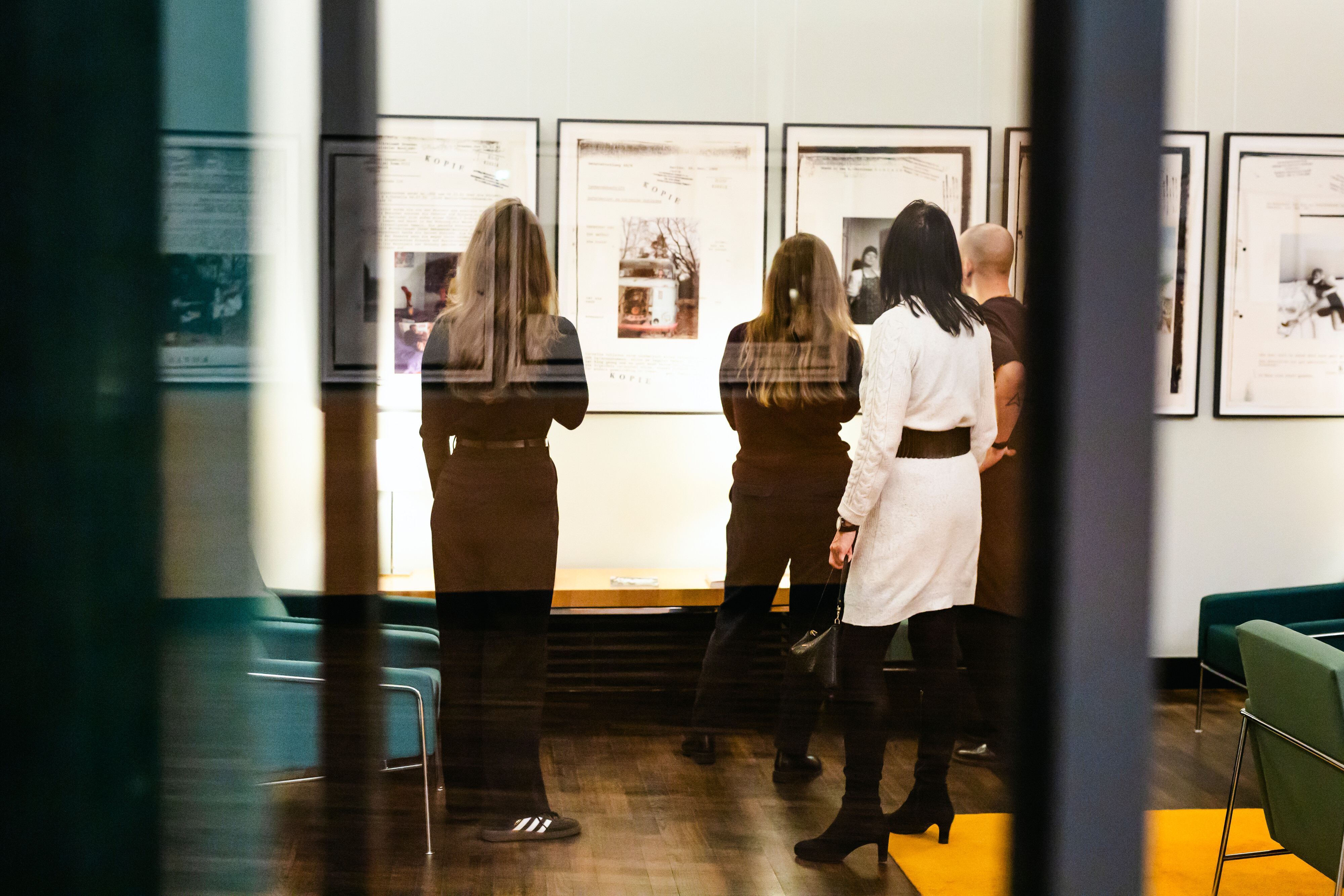 Vier Personen stehen in einer Kunstgalerie oder einem Museum und betrachten gerahmte Bilder oder Dokumente, die an einer Wand ausgestellt sind. Die Szene wird durch eine Glaswand betrachtet, die eine subtile Reflexion erzeugt.