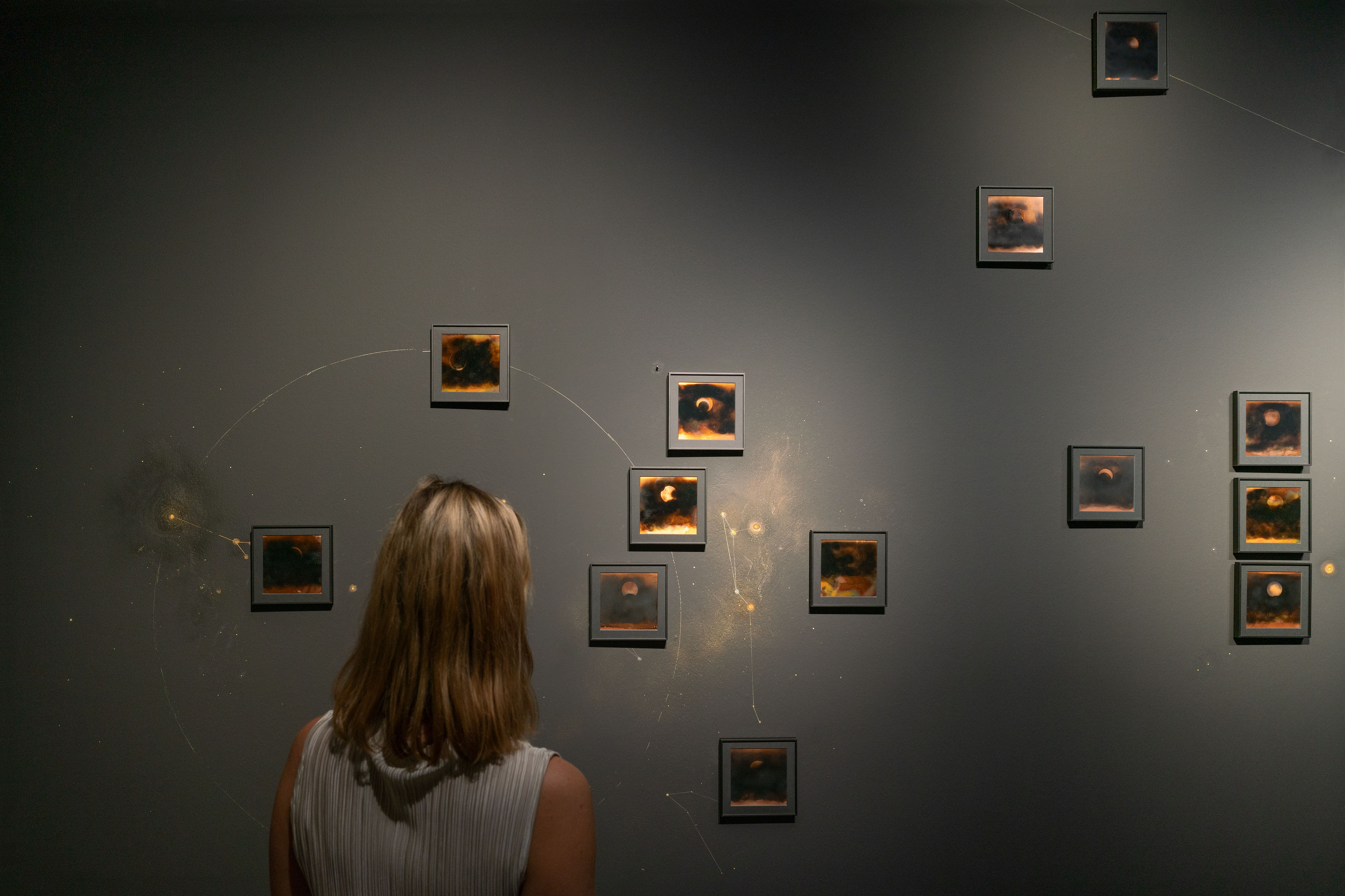 ine Frau mit blondem Haar betrachtet mehrere kleine, quadratische Kunstwerke an einer dunklen Wand, die Himmels- oder Naturphänomene zeigen, umgeben von skizzierten Linien und Punkten.