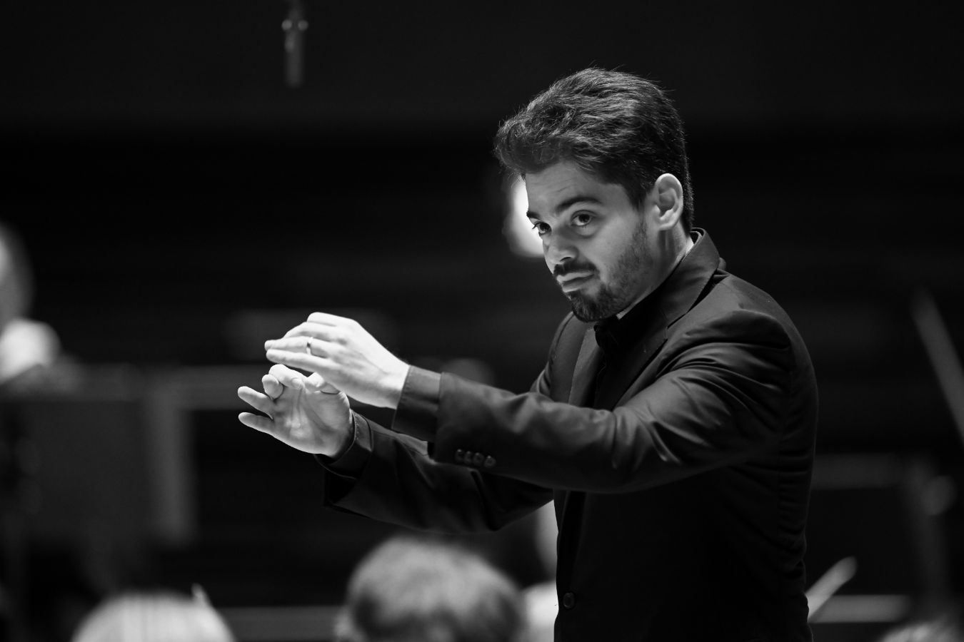 Lahav Shani in a dark suit conducts an orchestra, holding his hands mid-air with a focused expression. The background is blurred, emphasizing his gesture and facial concentration. The image is in black and white.